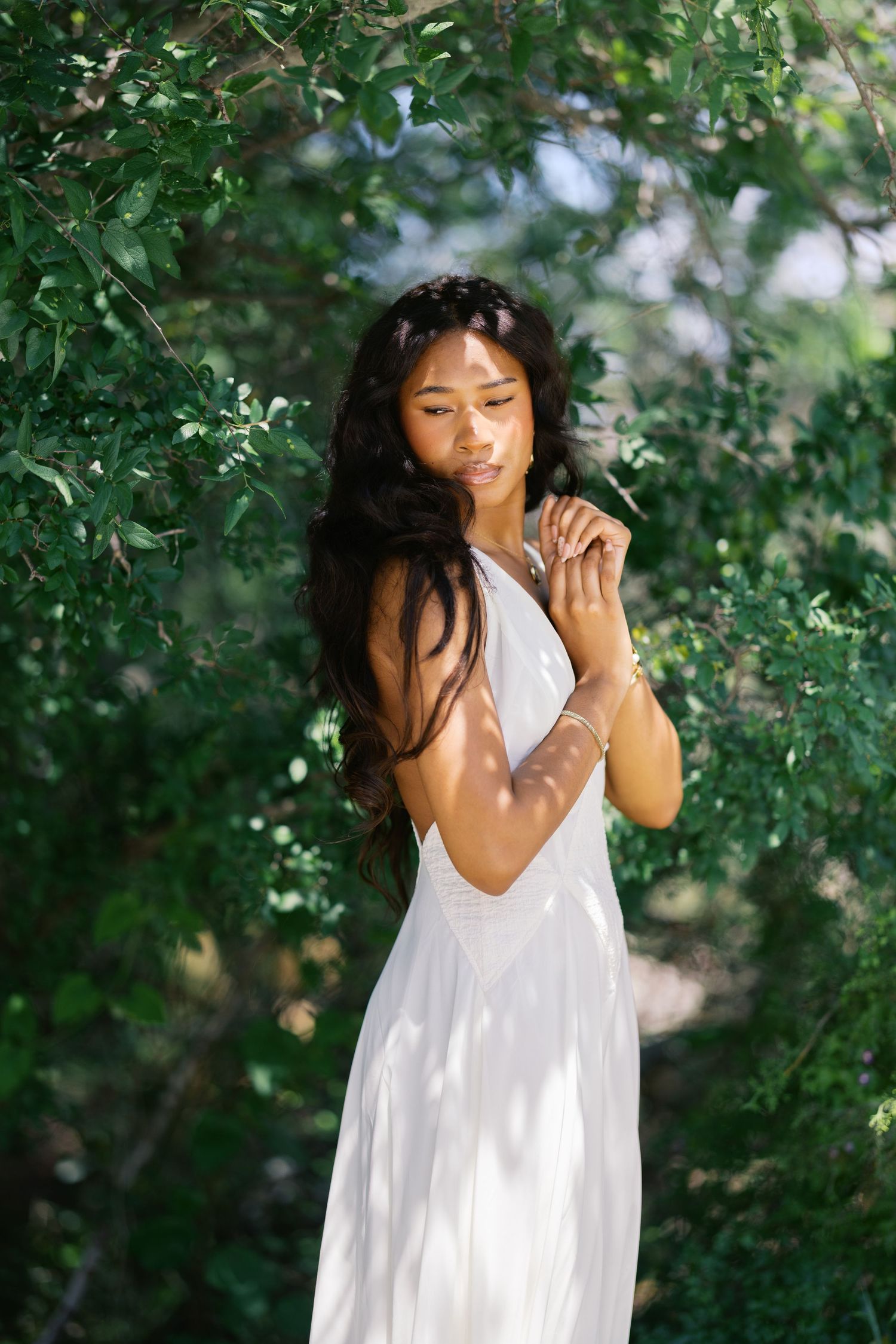 Young Black woman in flowing white dress standing among lush green tree branches outdoors.