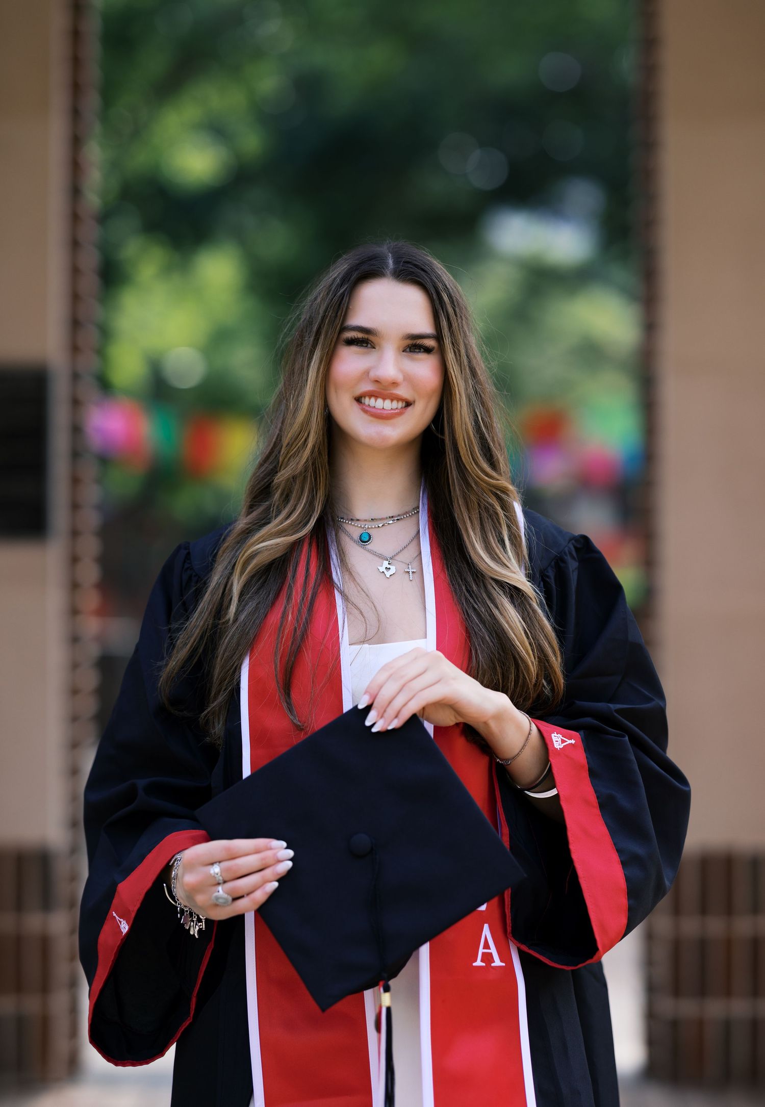 Smiling graduate in black cap and gown with red stole holding mortarboard outdoors.