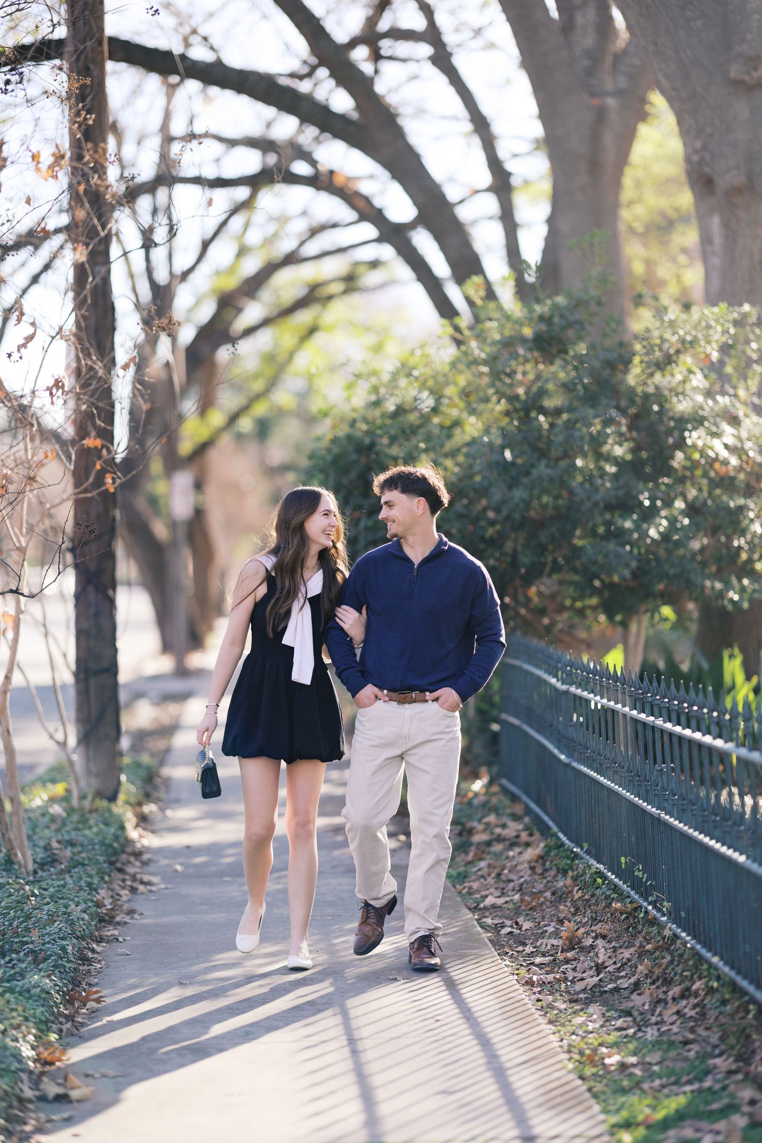 Couple walking together on a sunny tree-lined sidewalk, woman in black dress, man in navy sweater and khakis.