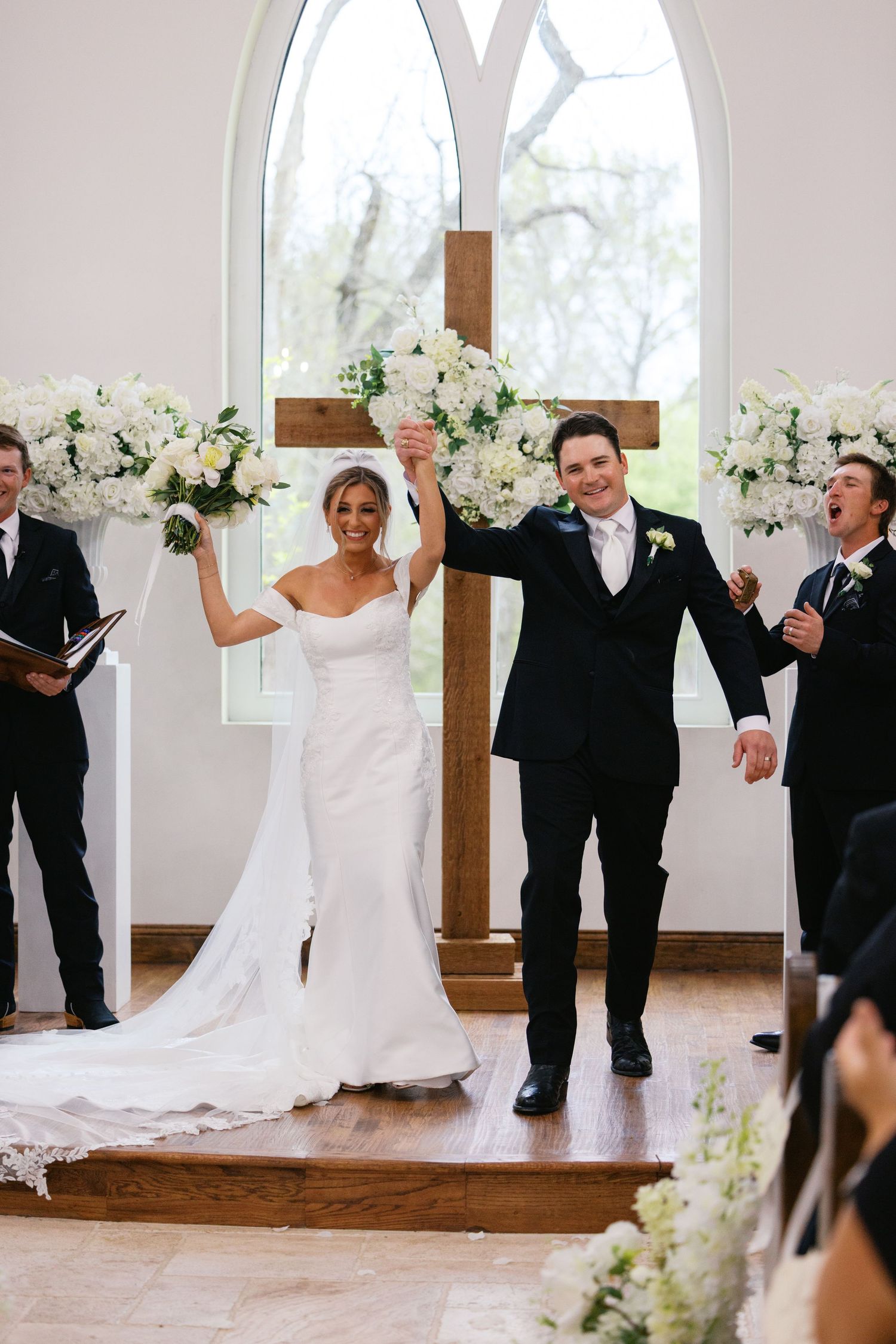 Joyful newlyweds raise hands at altar adorned with white floral arrangements and wooden cross after ceremony.
