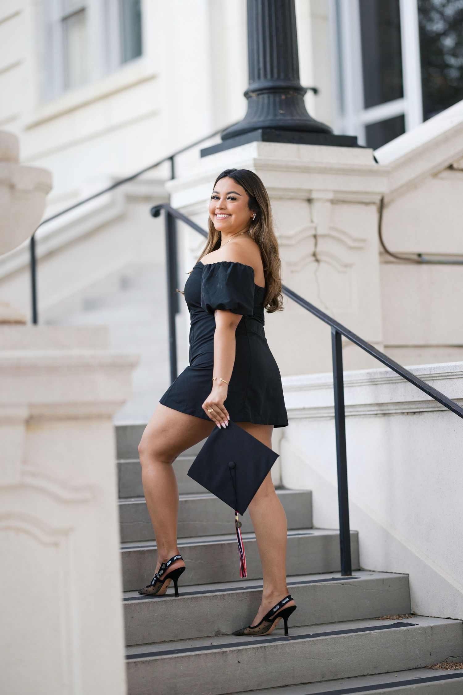 Young woman in black off-shoulder dress holding graduation cap on university steps, smiling confidently.
