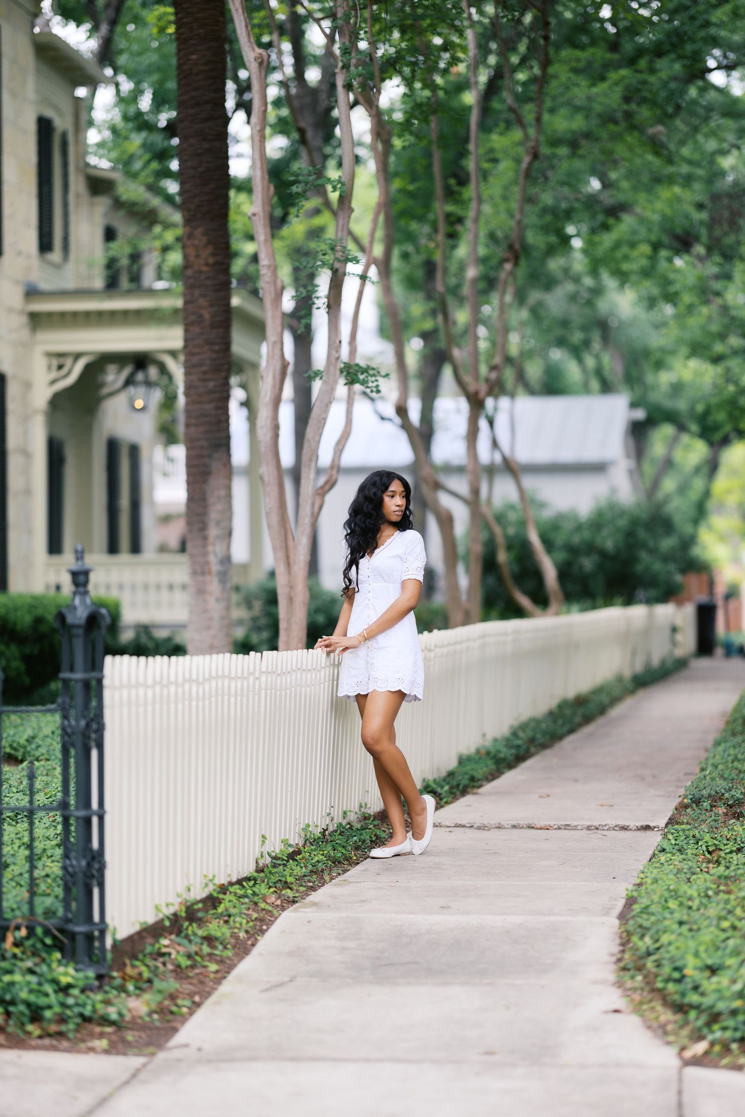 Young Black woman in white mini dress leans against white picket fence on tree-lined suburban street.