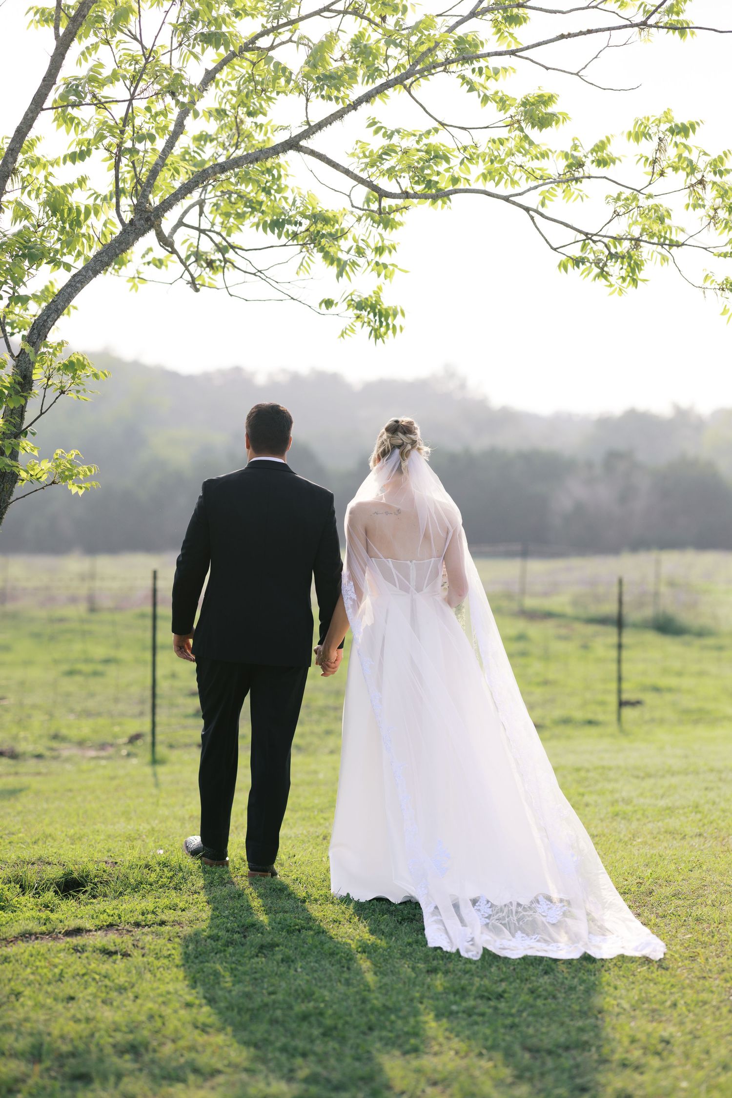 Bride and groom walk hand in hand across green field with wedding veil trailing.
