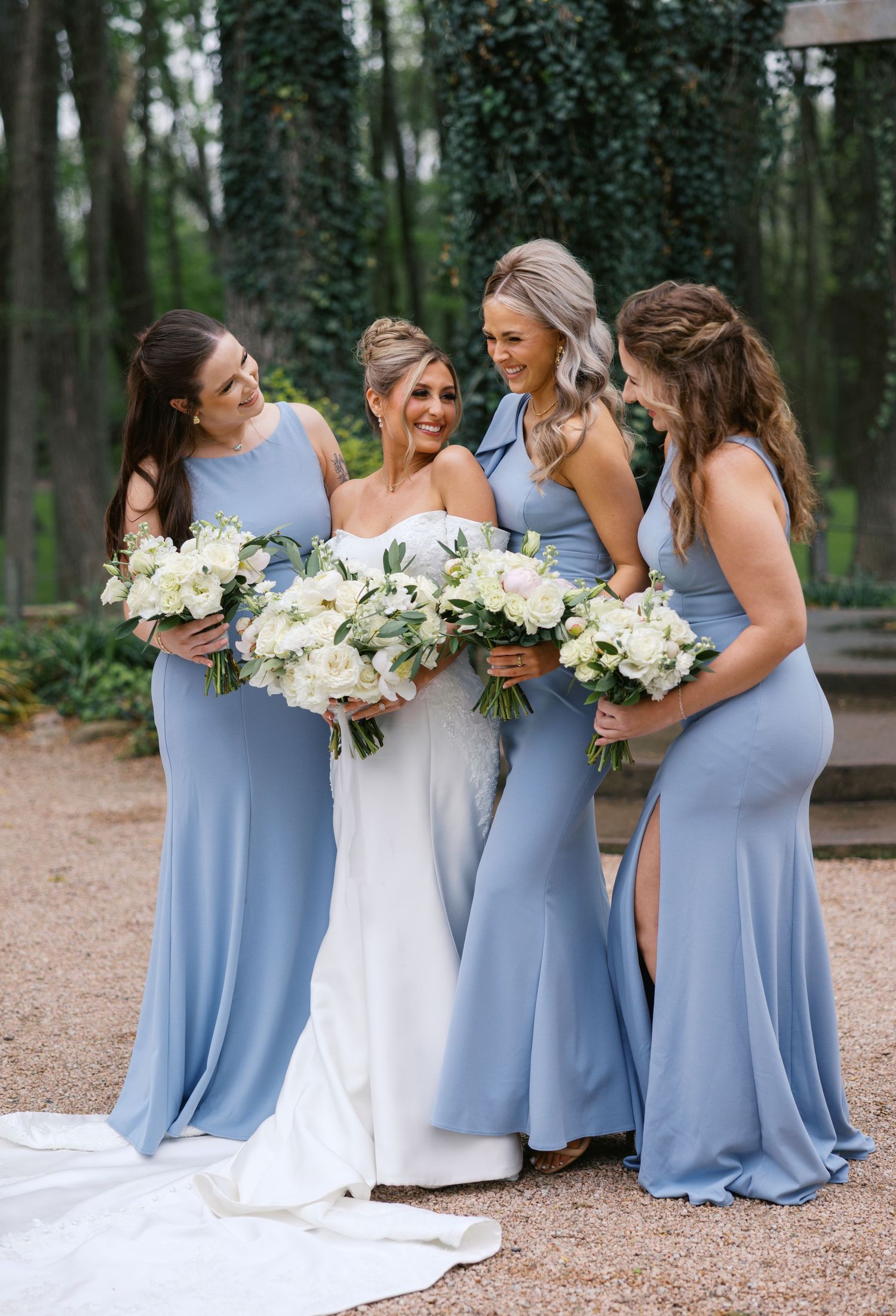Bride and three bridesmaids in dusty blue gowns laugh together while holding white floral bouquets outdoors.