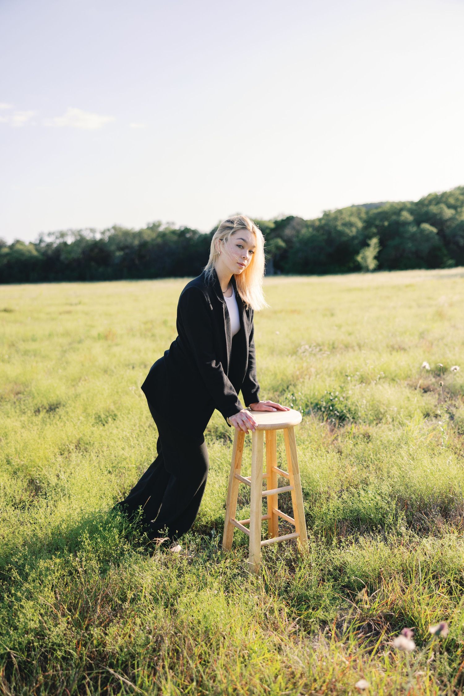 Figure in black attire leaning on wooden stool in grassy field during golden hour.