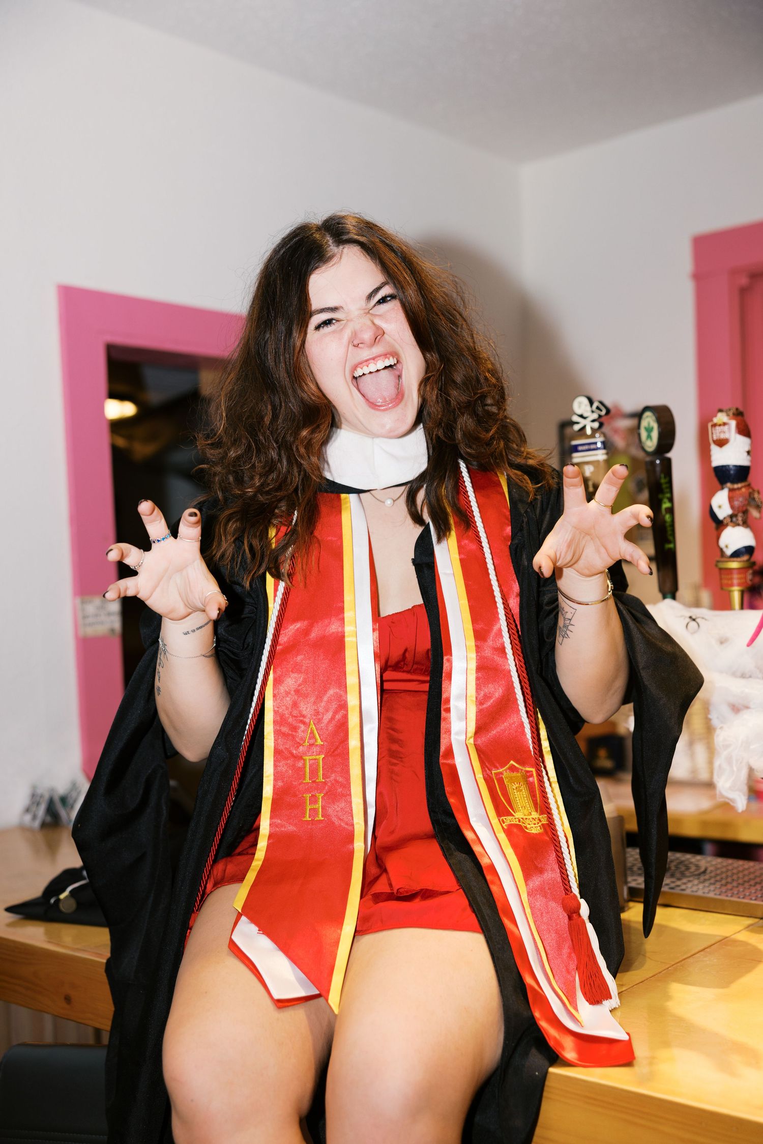Excited graduate in red stoles and black gown celebrating indoors.