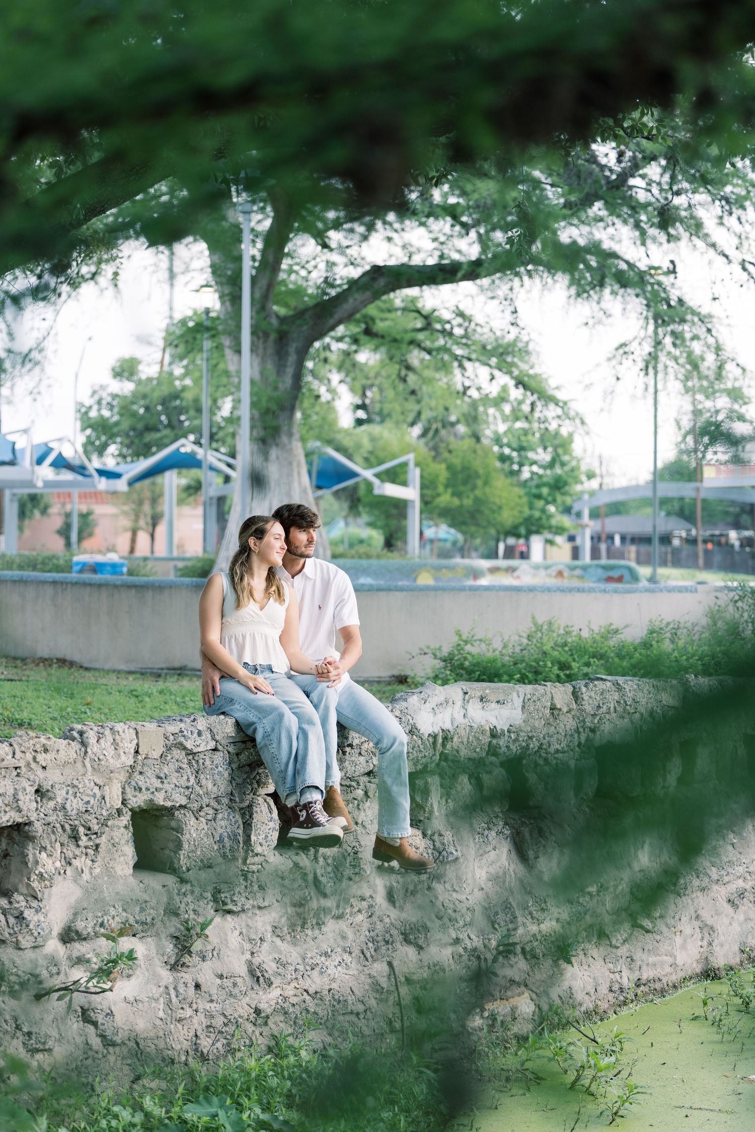 A couple sits casually on a stone wall surrounded by greenery near water.