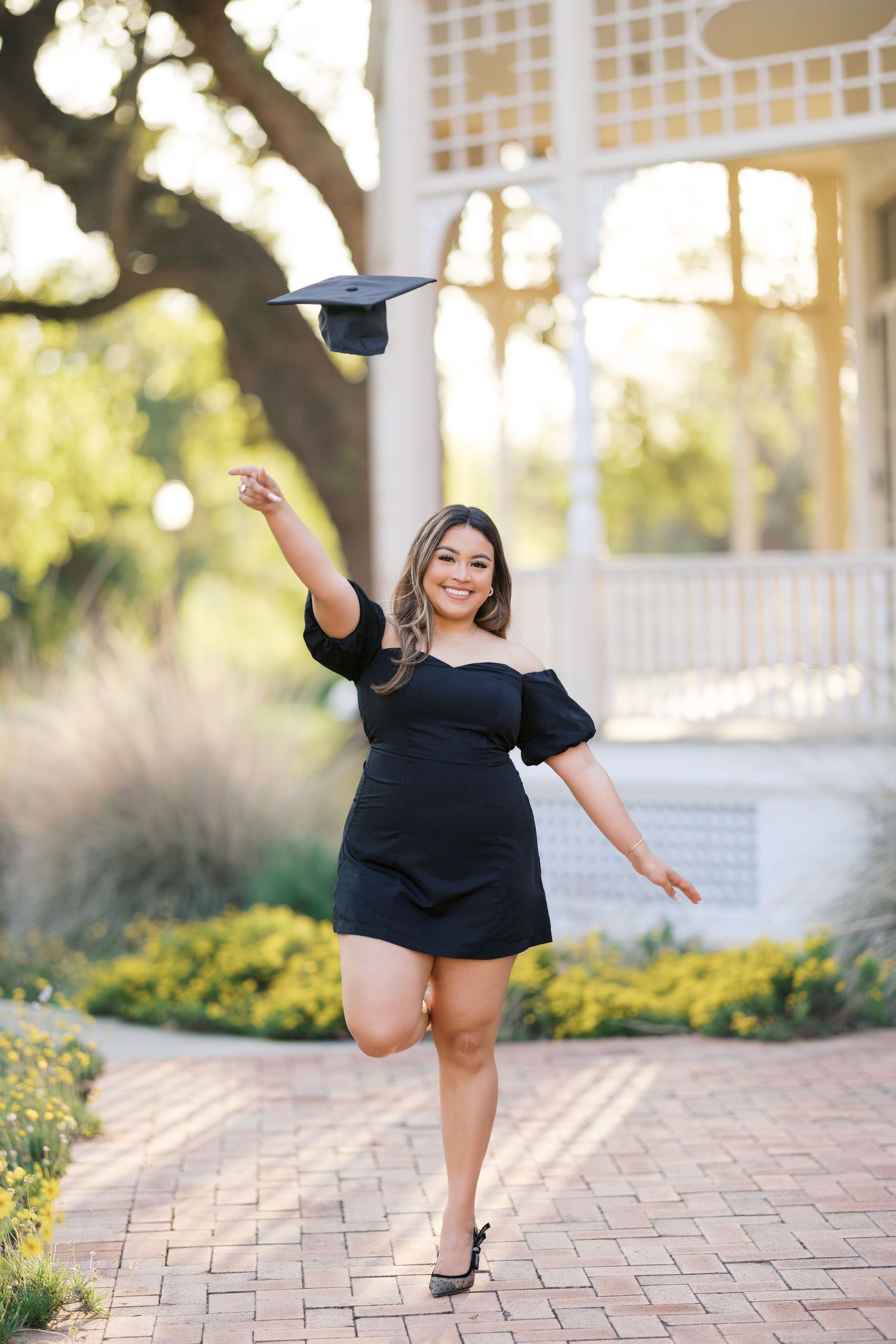 A smiling graduate in a black dress tosses her cap in the air outdoors near a gazebo.
