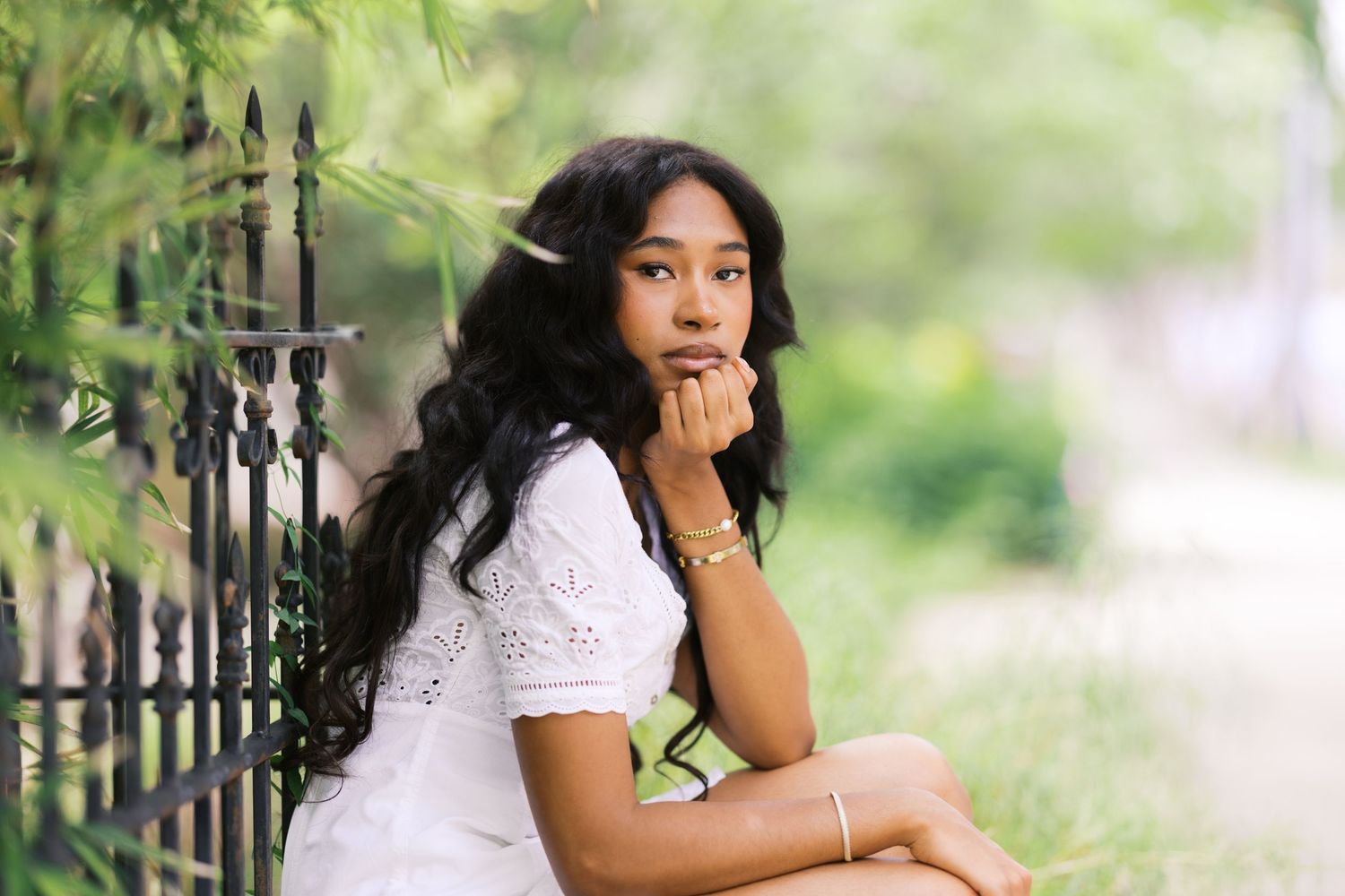 Young Black woman in white lace dress sitting by iron fence outdoors, resting chin on hand, surrounded by lush greenery.