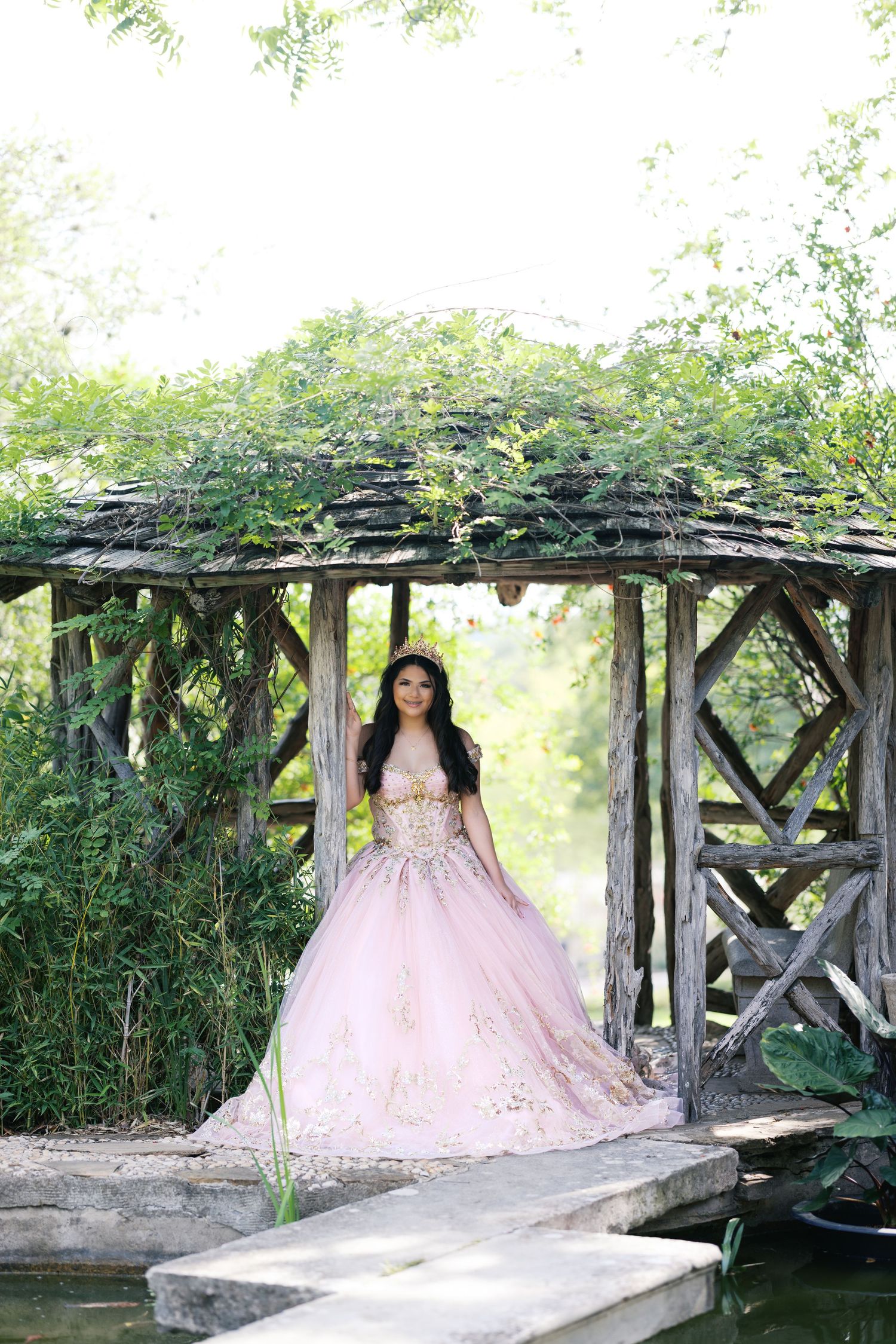Young woman in pink gold quinceañera ball gown poses under rustic wooden gazebo covered in lush green vines.