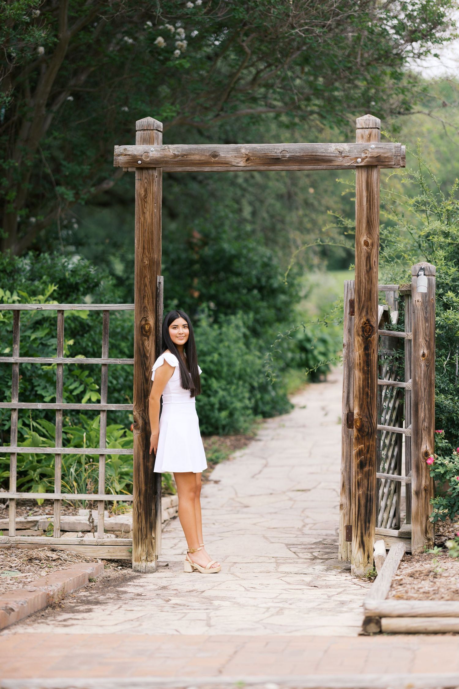 Young woman in white dress standing under rustic wooden garden gate surrounded by lush greenery.
