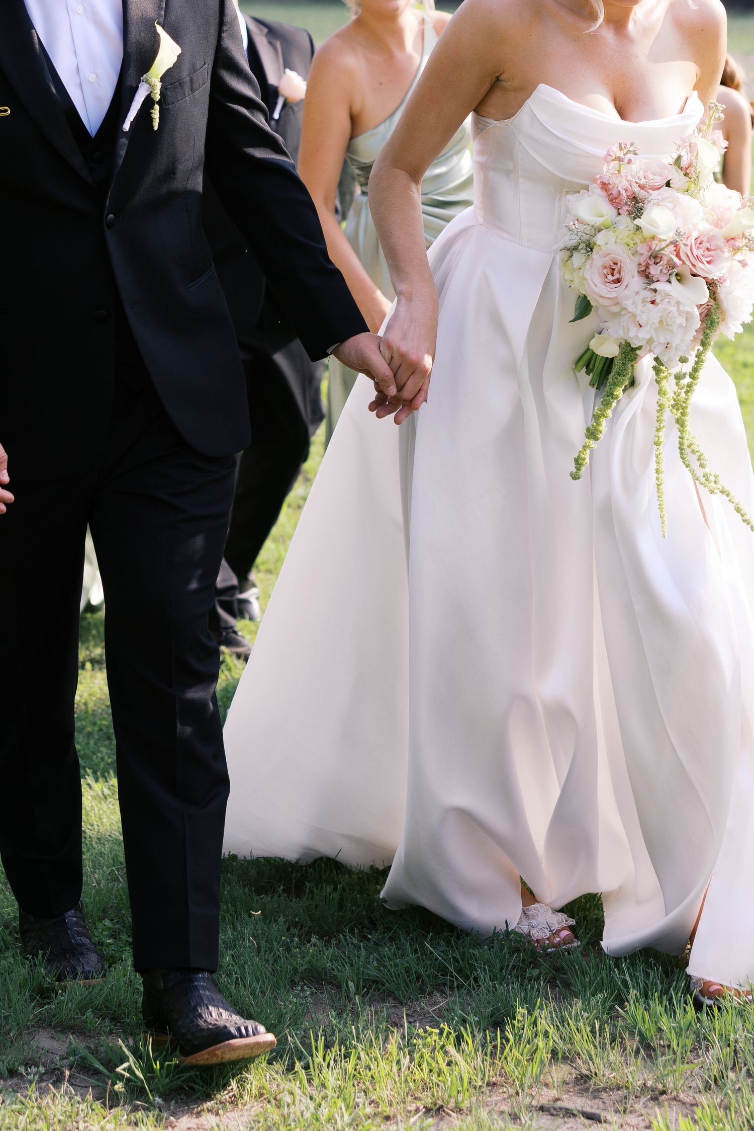 Bride in white strapless gown holding pink floral bouquet walks hand in hand with groom in black suit on grass.