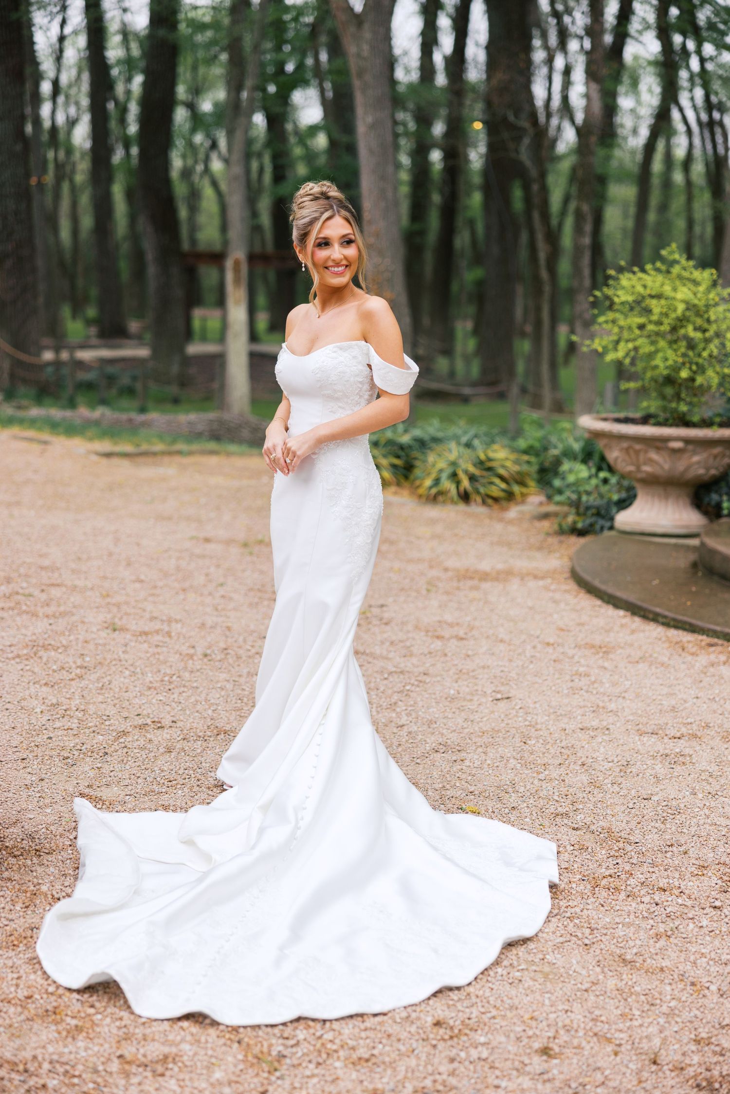 Elegant bride poses in an off-shoulder white mermaid wedding gown with a long train on a garden gravel path.