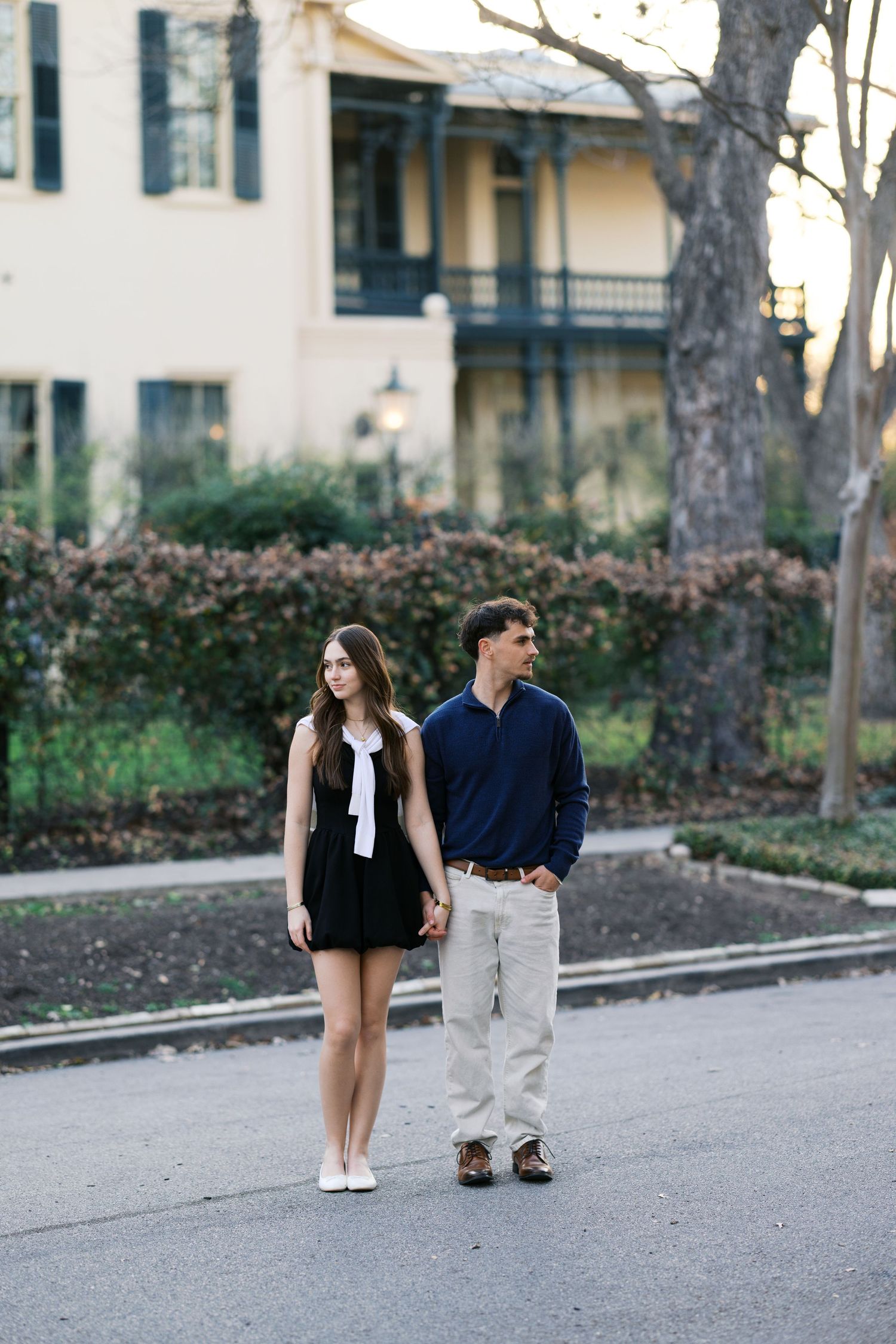 Couple holding hands walking on a quiet street with a yellow historic building and manicured hedges in background.