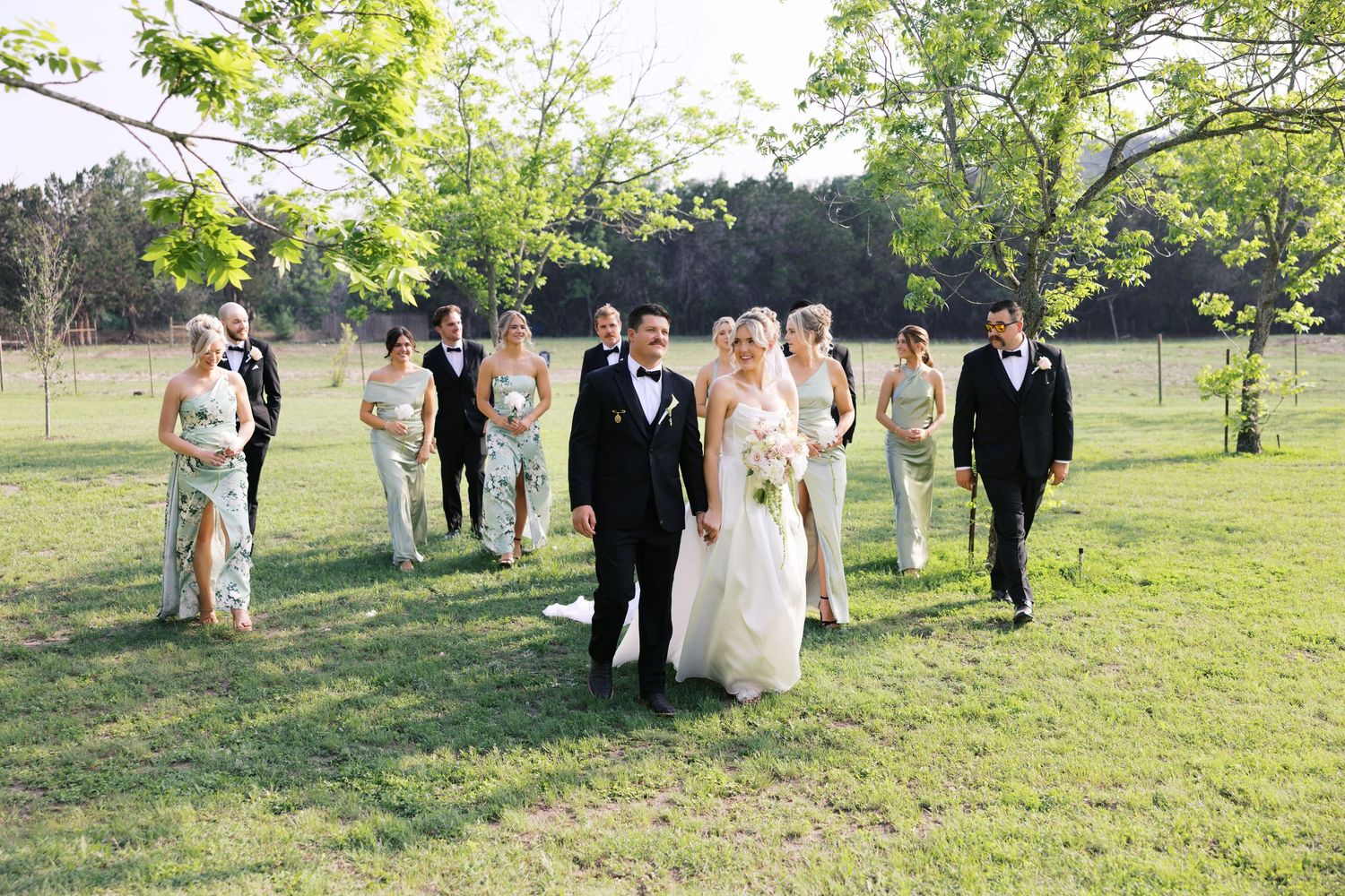 Bride and groom walking with wedding party on green lawn, bridesmaids in sage dresses and groomsmen in black tuxedos.