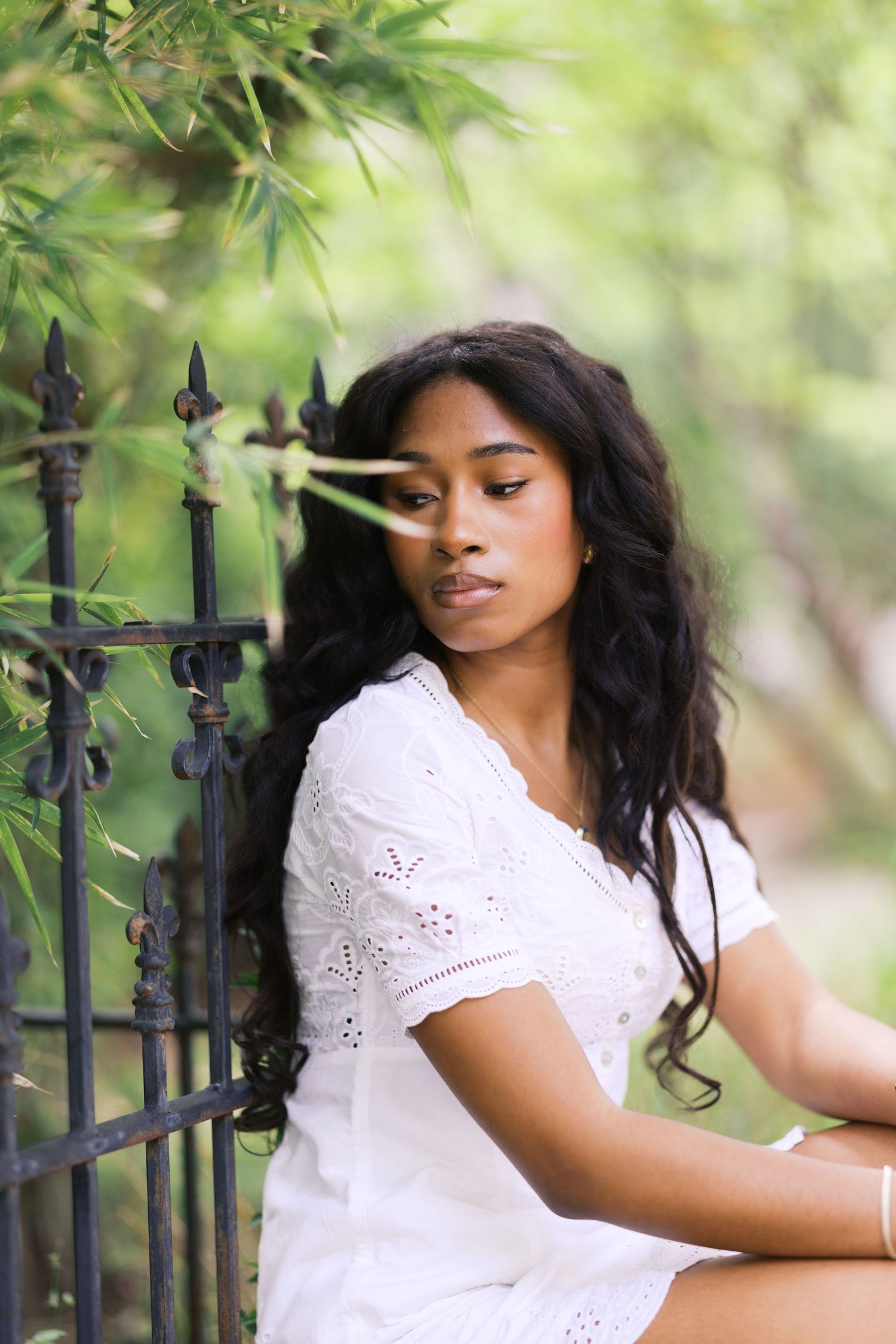 Young Black woman in white eyelet dress sits pensively by iron fence amid lush green foliage.