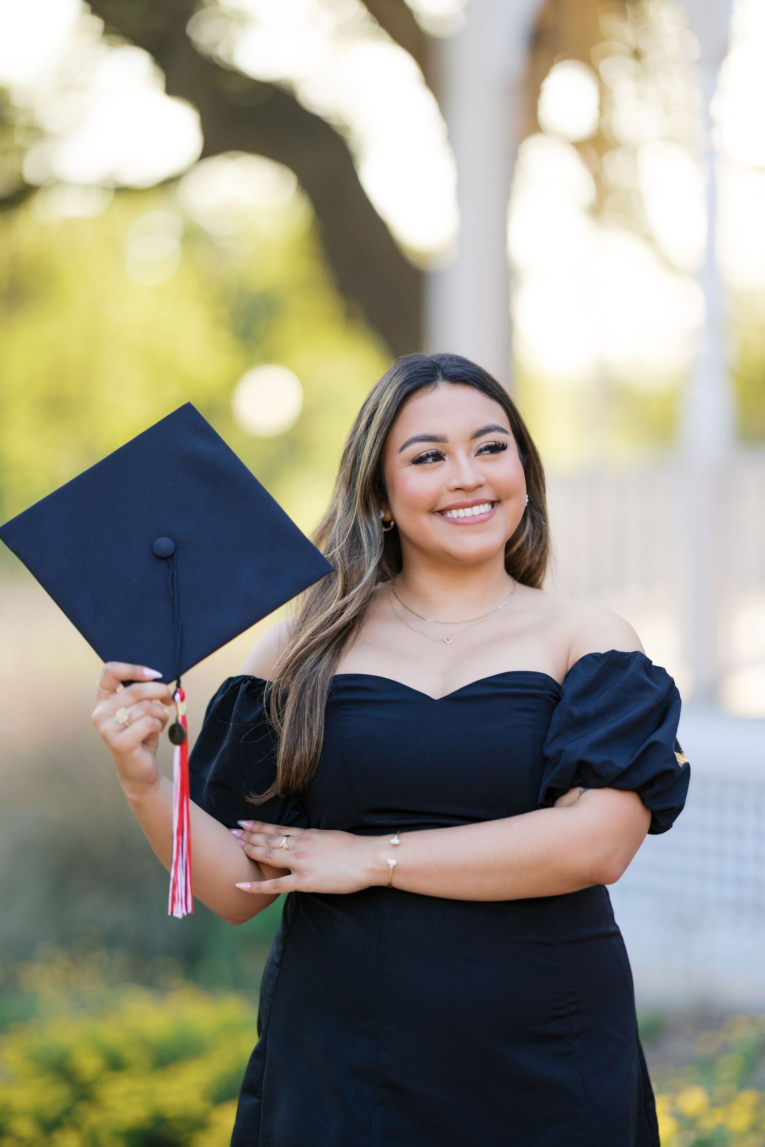Happy graduate in black off-shoulder dress holds navy mortarboard cap outdoors.