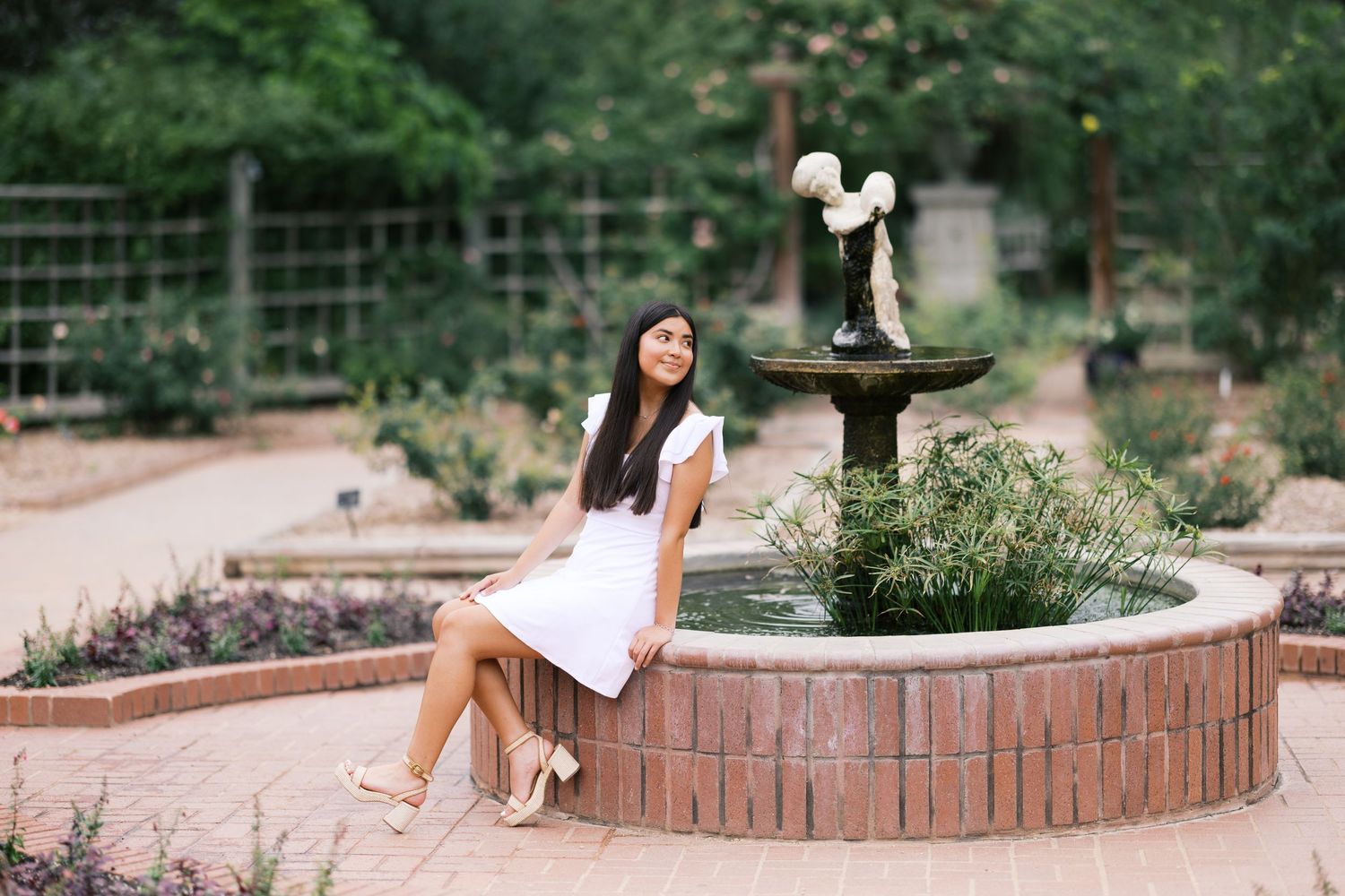 Young woman in white dress sitting on brick fountain edge in a lush garden setting.