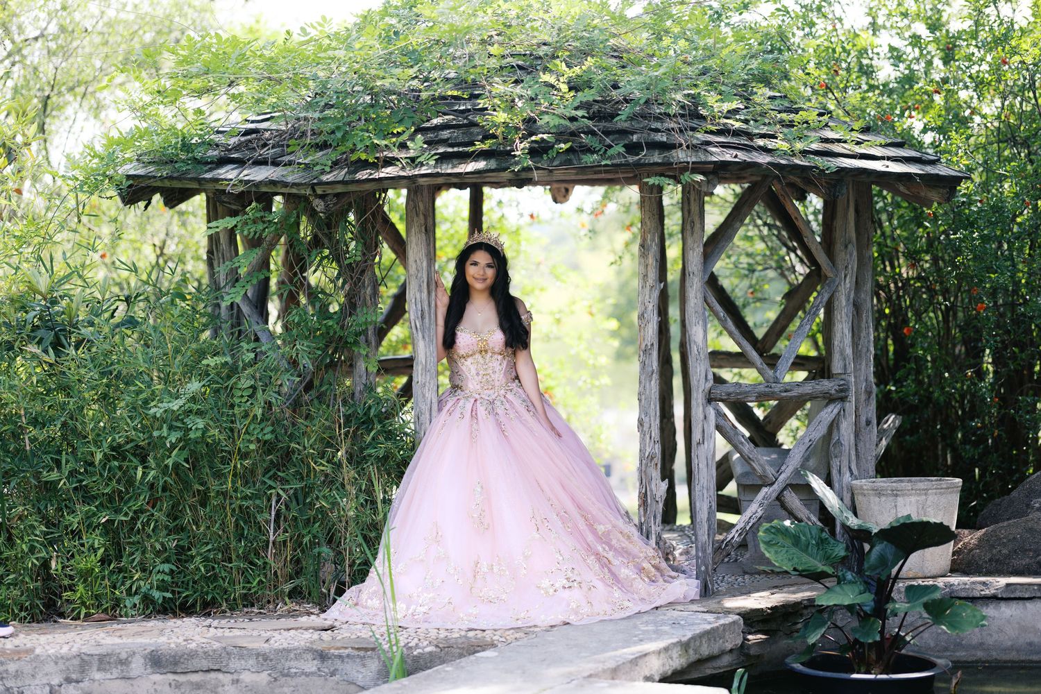 Young woman in pink ball gown poses under rustic wooden gazebo surrounded by lush green garden foliage.