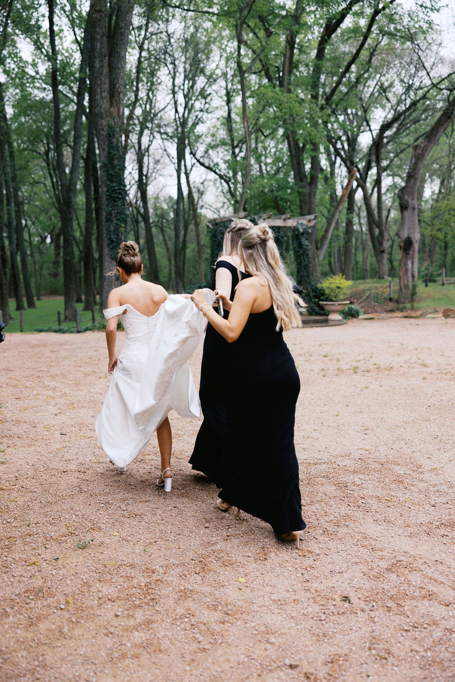 Bride in white wedding gown walking through wooded garden path assisted by bridesmaid in black.