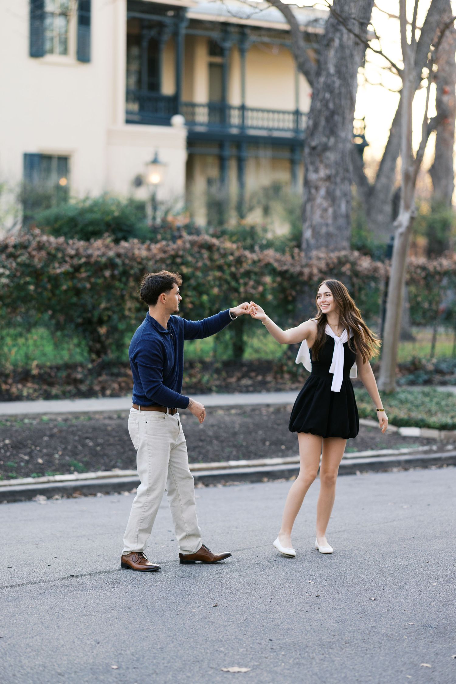 A stylish couple holds hands and dances on a tree-lined street, both dressed in navy and black outfits.