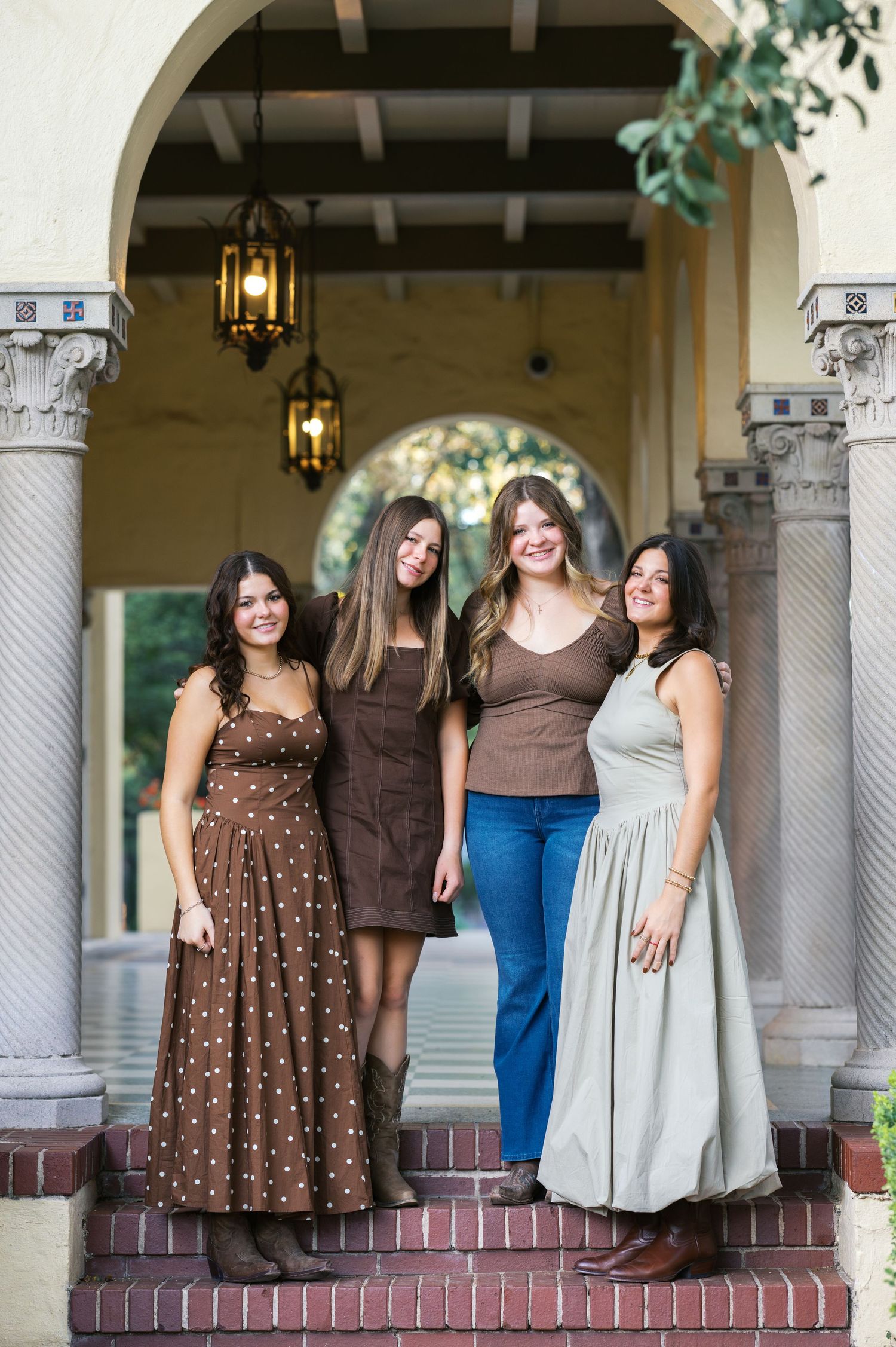 Four women in coordinated brown and neutral outfits pose on brick steps under an archway.