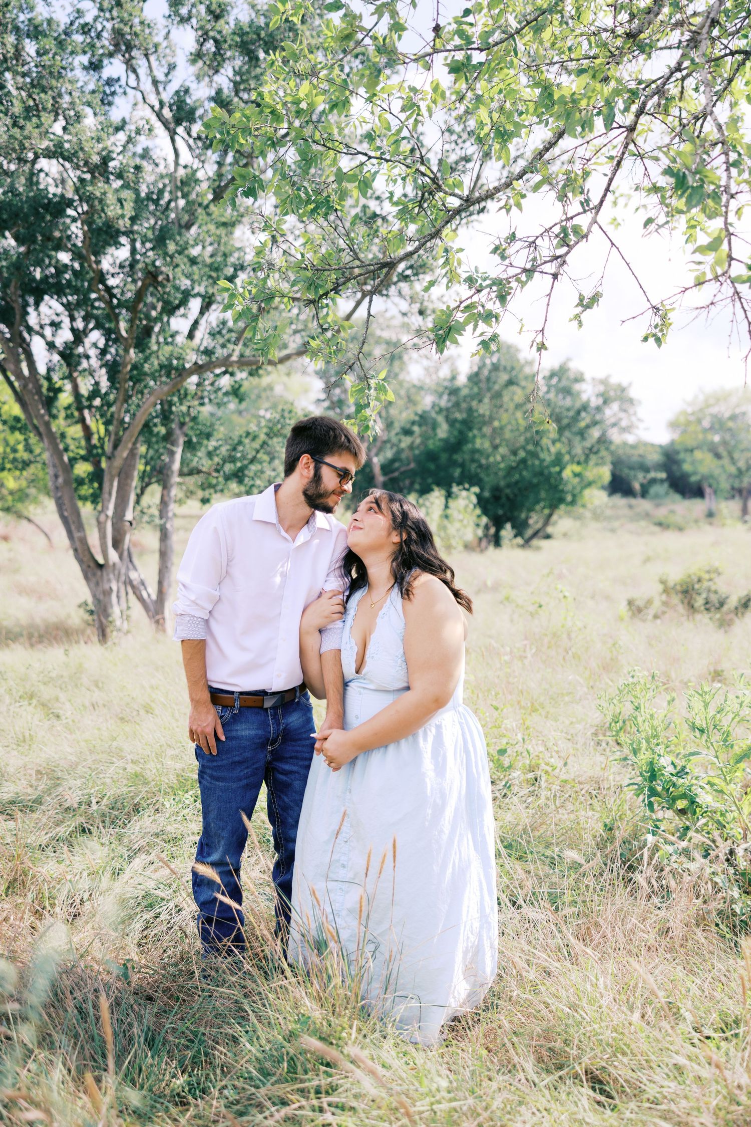 A romantic couple embraces in a sunlit meadow with trees in the background.
