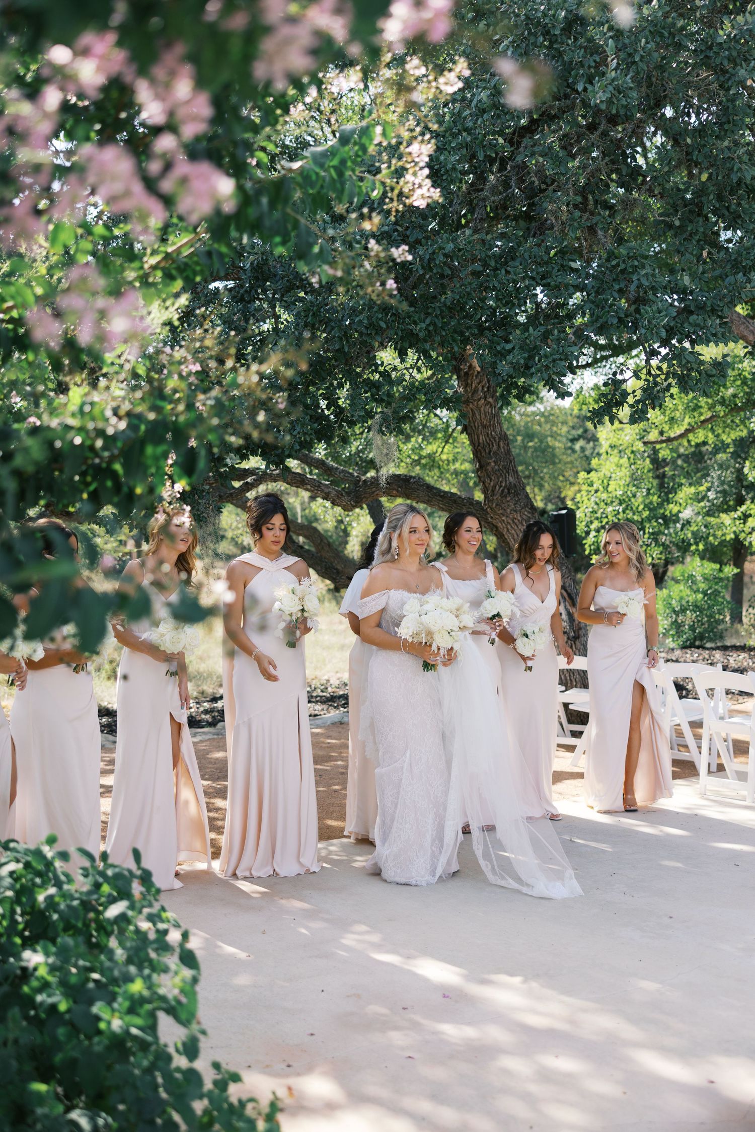 Bridal party in elegant white and blush dresses pose together under flowering trees at an outdoor wedding venue.