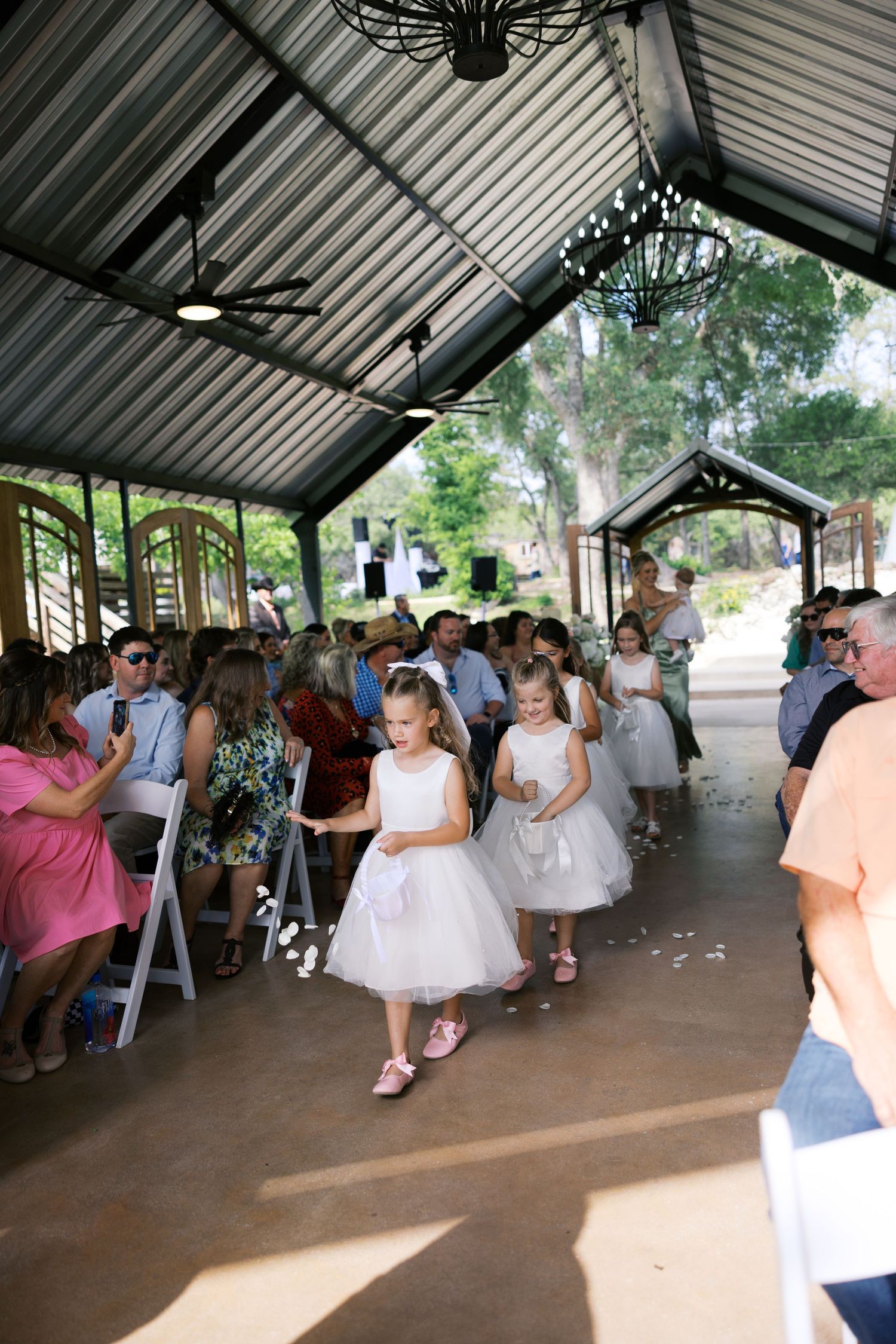 Two flower girls in white dresses walk down the aisle at an outdoor wedding ceremony under a pavilion.