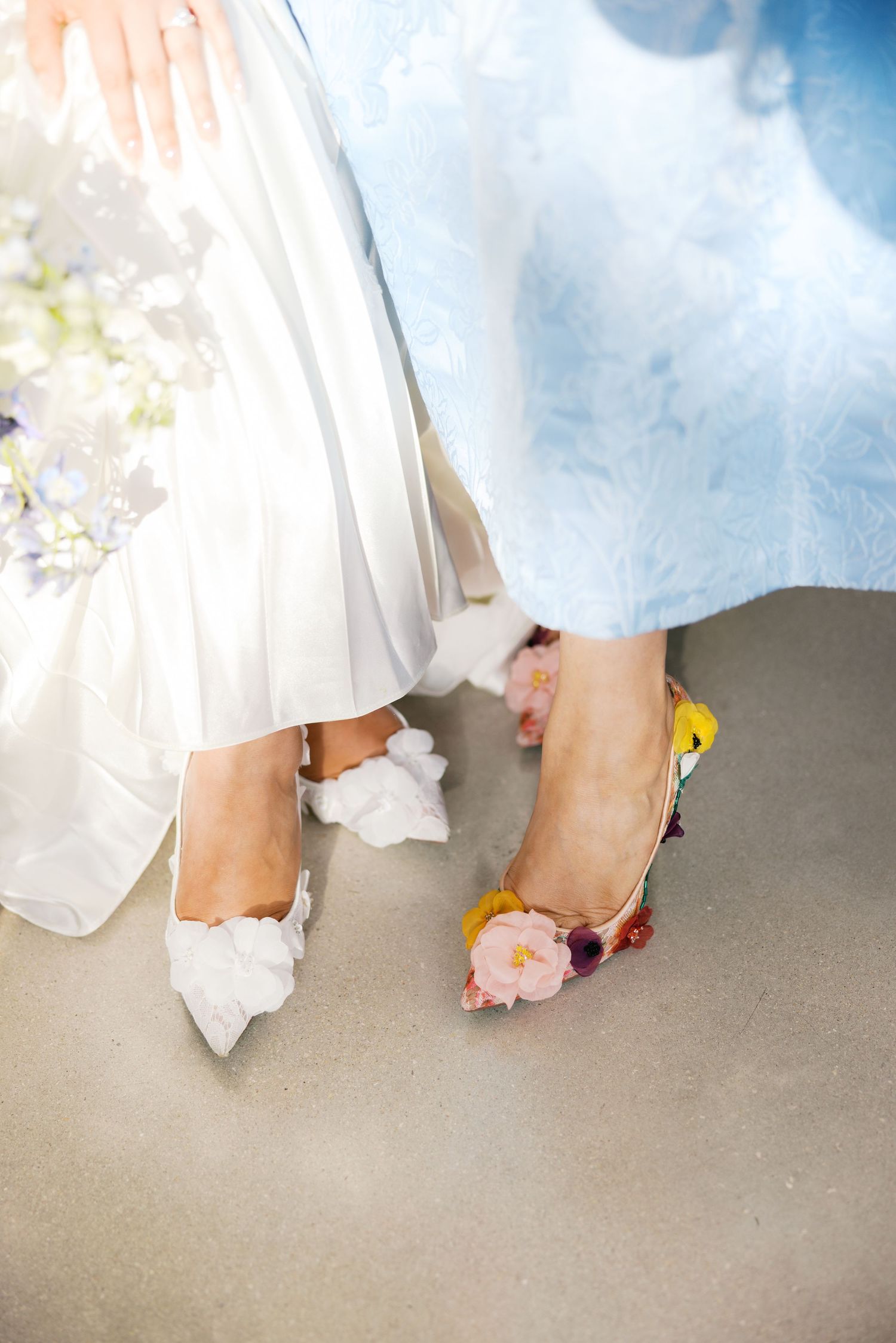 Close-up of two women's feet wearing elegant floral heels, one in white and one in blue dress.