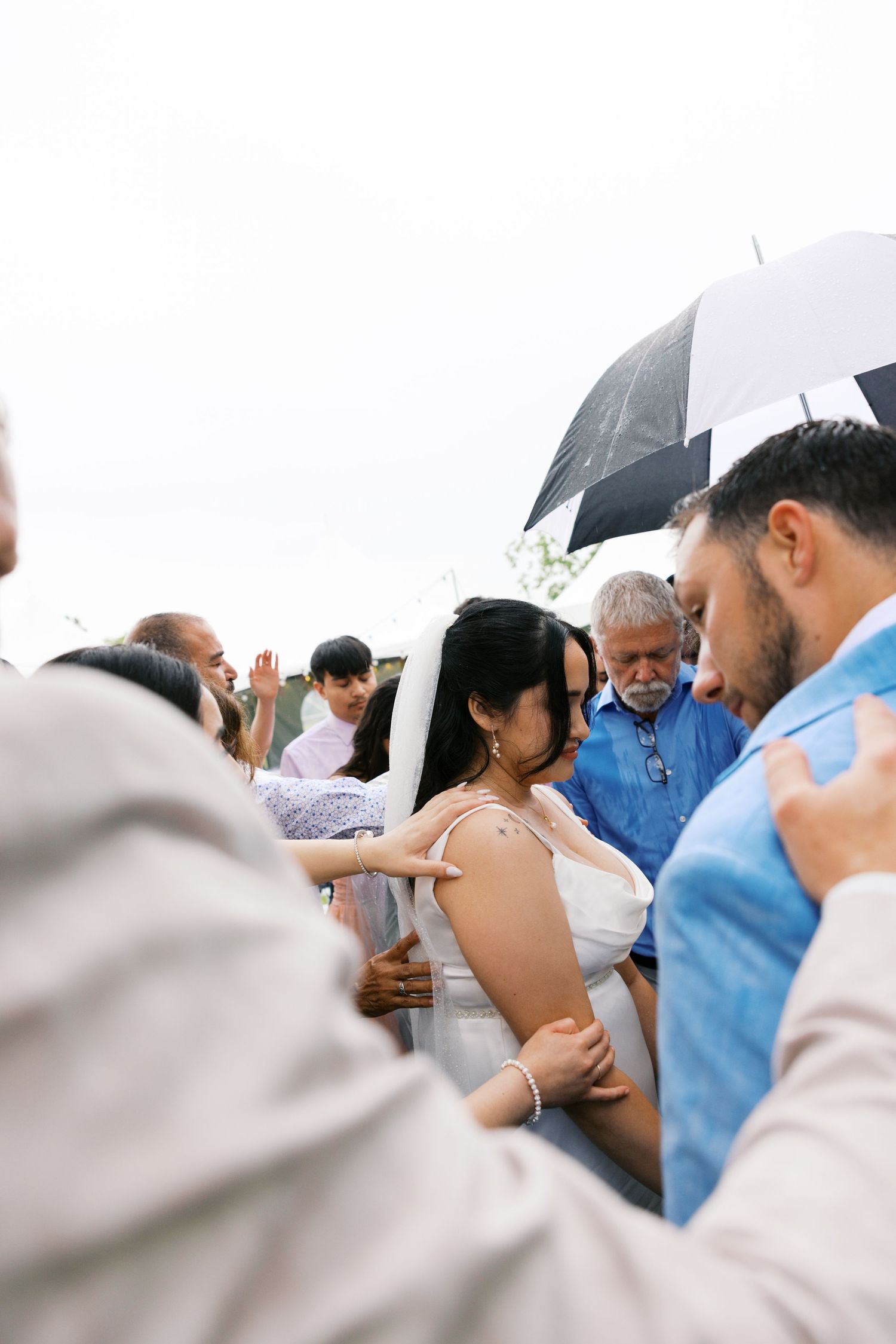 Wedding guests surround bride in white dress during outdoor ceremony blessing under cloudy sky.