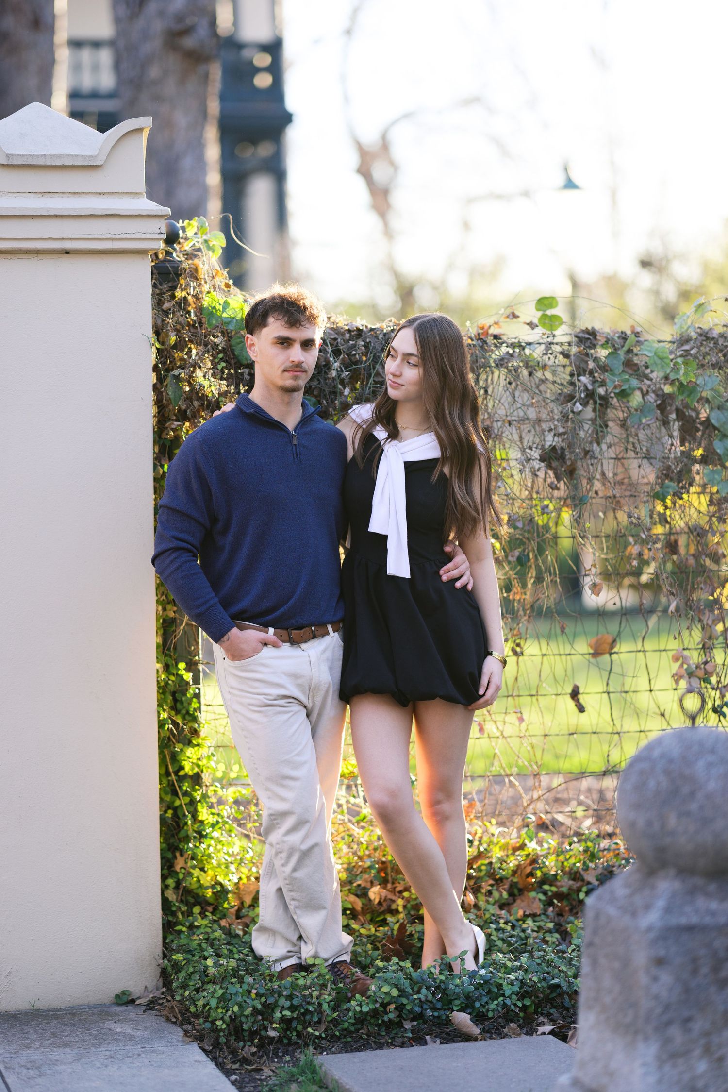 Stylish couple posing by a vine-covered gate, woman in black dress with white bow, man in navy sweater.
