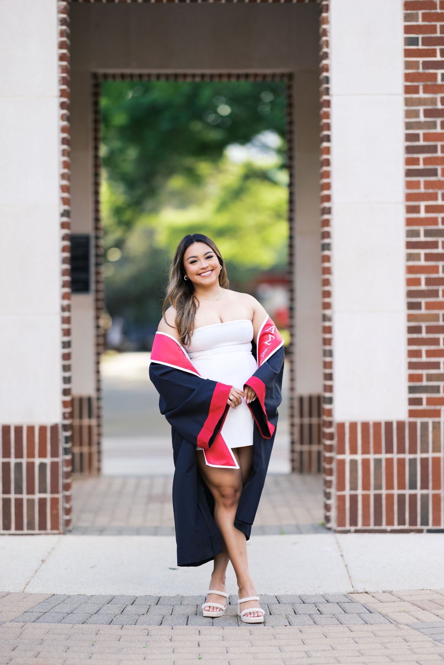 Young woman in white dress holding graduation robe poses under brick archway on college campus.