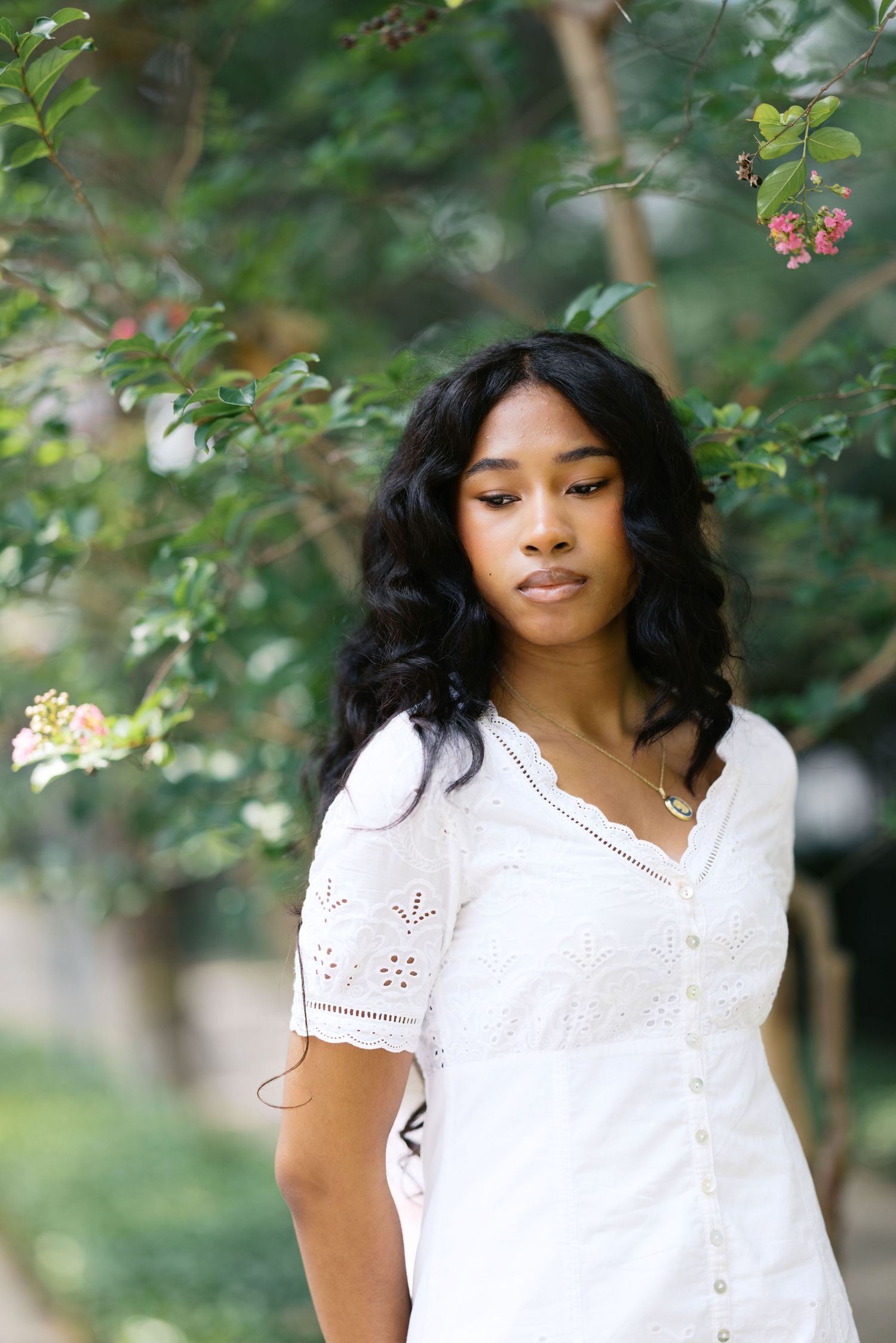 Young Black woman in white eyelet dress standing near blooming pink flowers on tree.