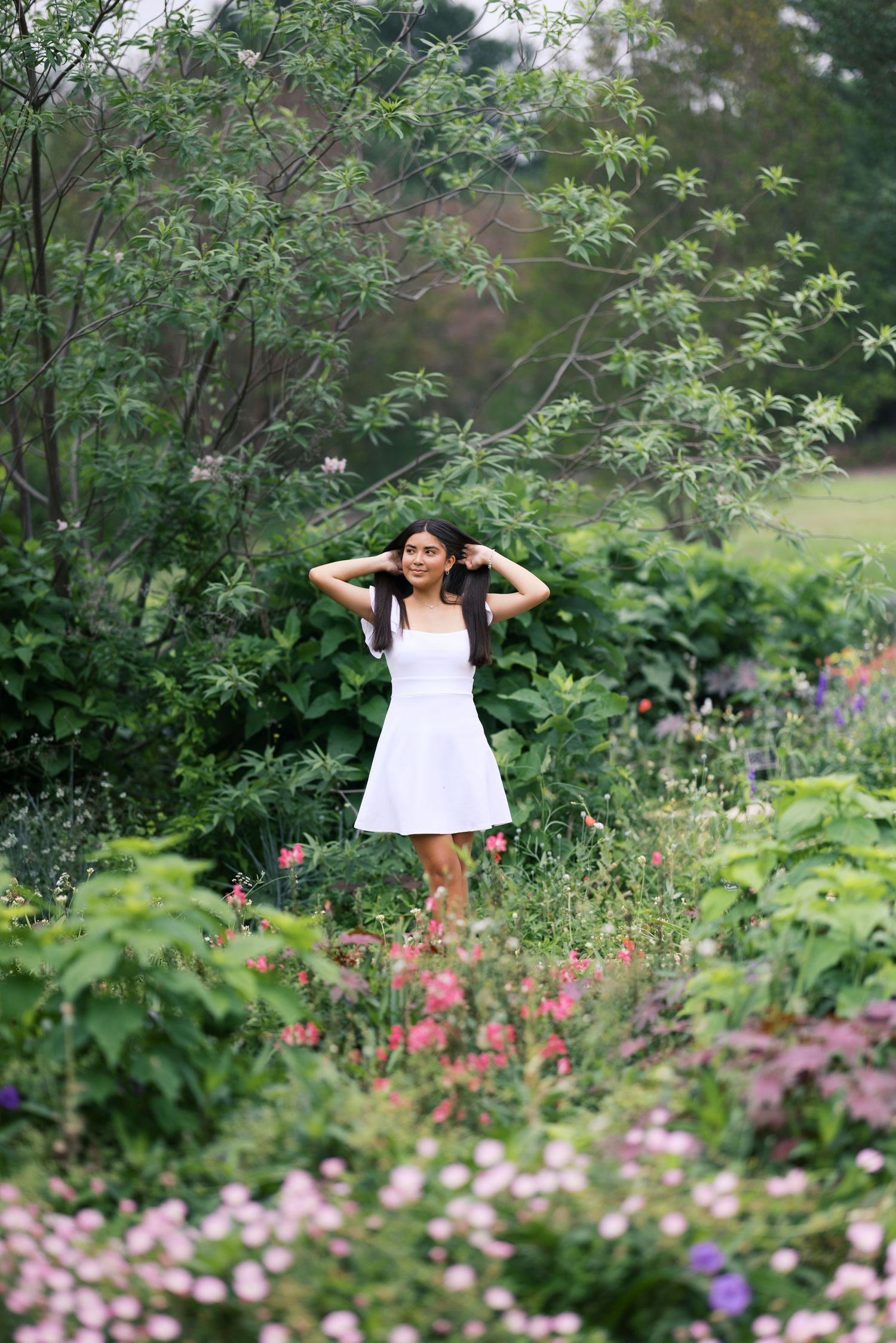 Young woman in white dress standing among colorful garden flowers and lush green trees, hands in hair.