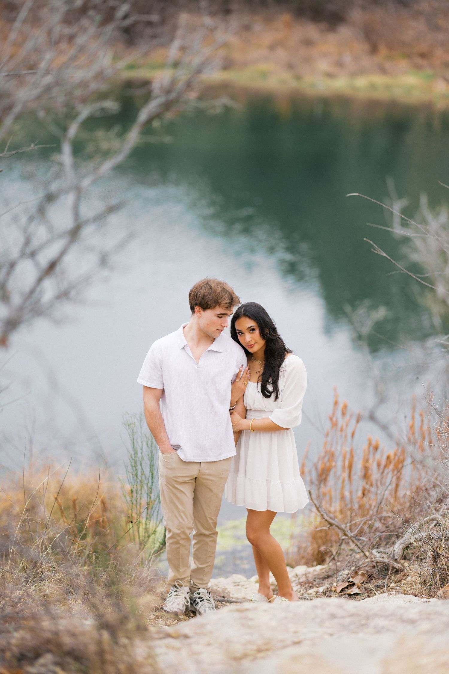Couple in white outfits sharing an intimate moment by a serene lake with dry grass in the foreground.