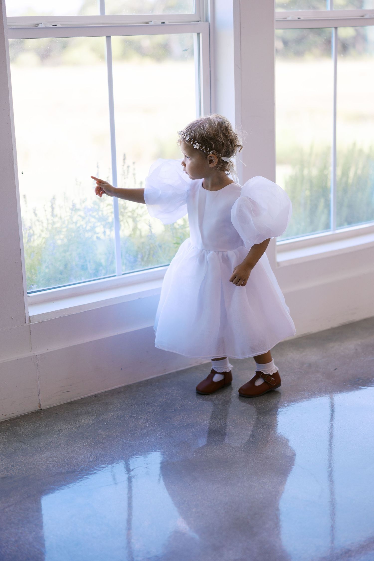 Young flower girl in puffy white dress and mary janes points at window.