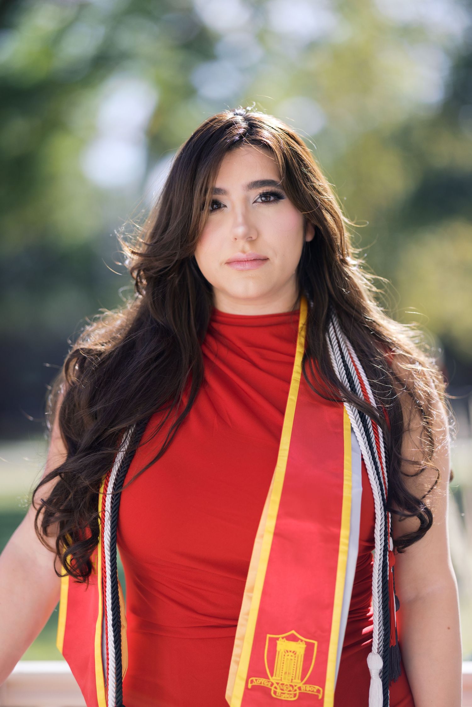 Red graduation gown with yellow and white sash shown against blurred outdoor background.