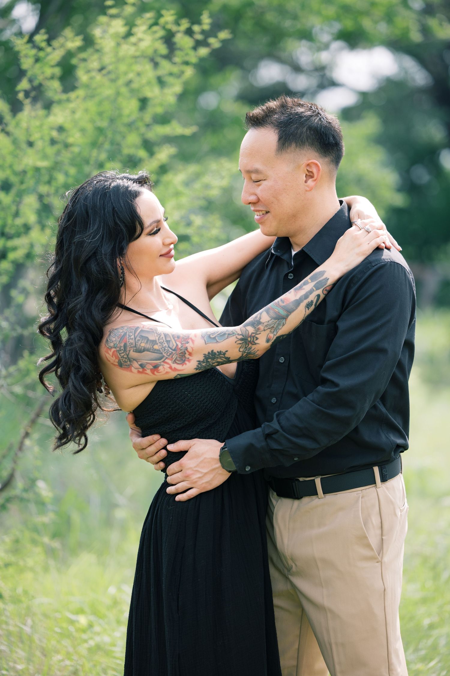 Couple in black attire embracing outdoors with visible tattoo sleeve and natural greenery.