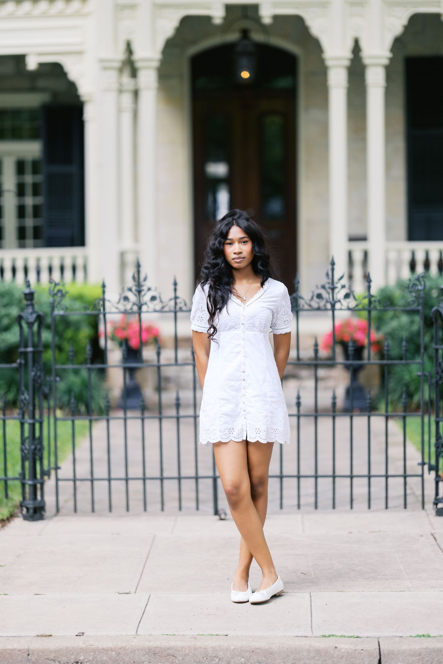 Young woman in white eyelet mini dress and flats standing on sidewalk before ornate iron fence and historic home.