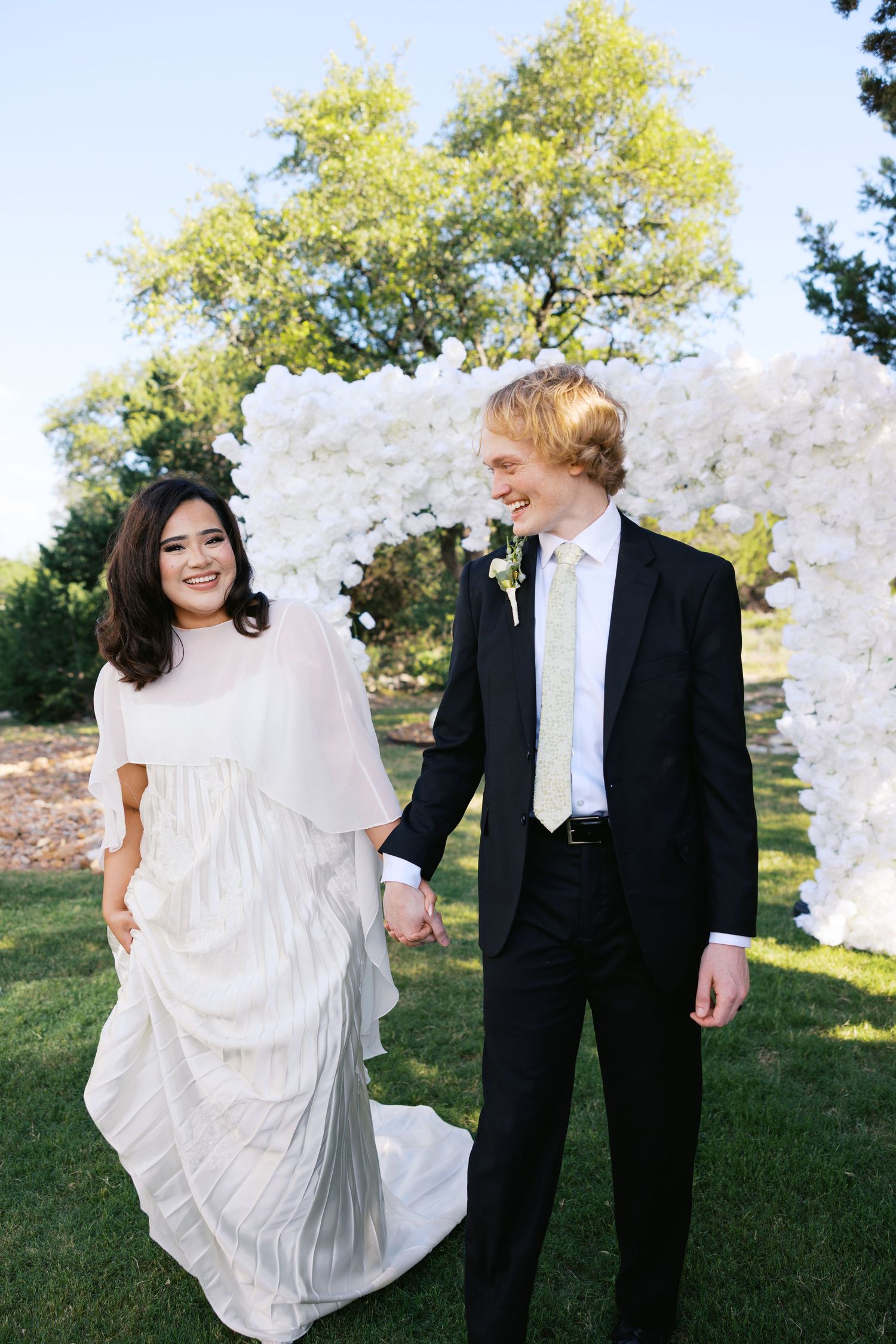 A smiling bride and groom hold hands outdoors near a white floral arch on their wedding day.