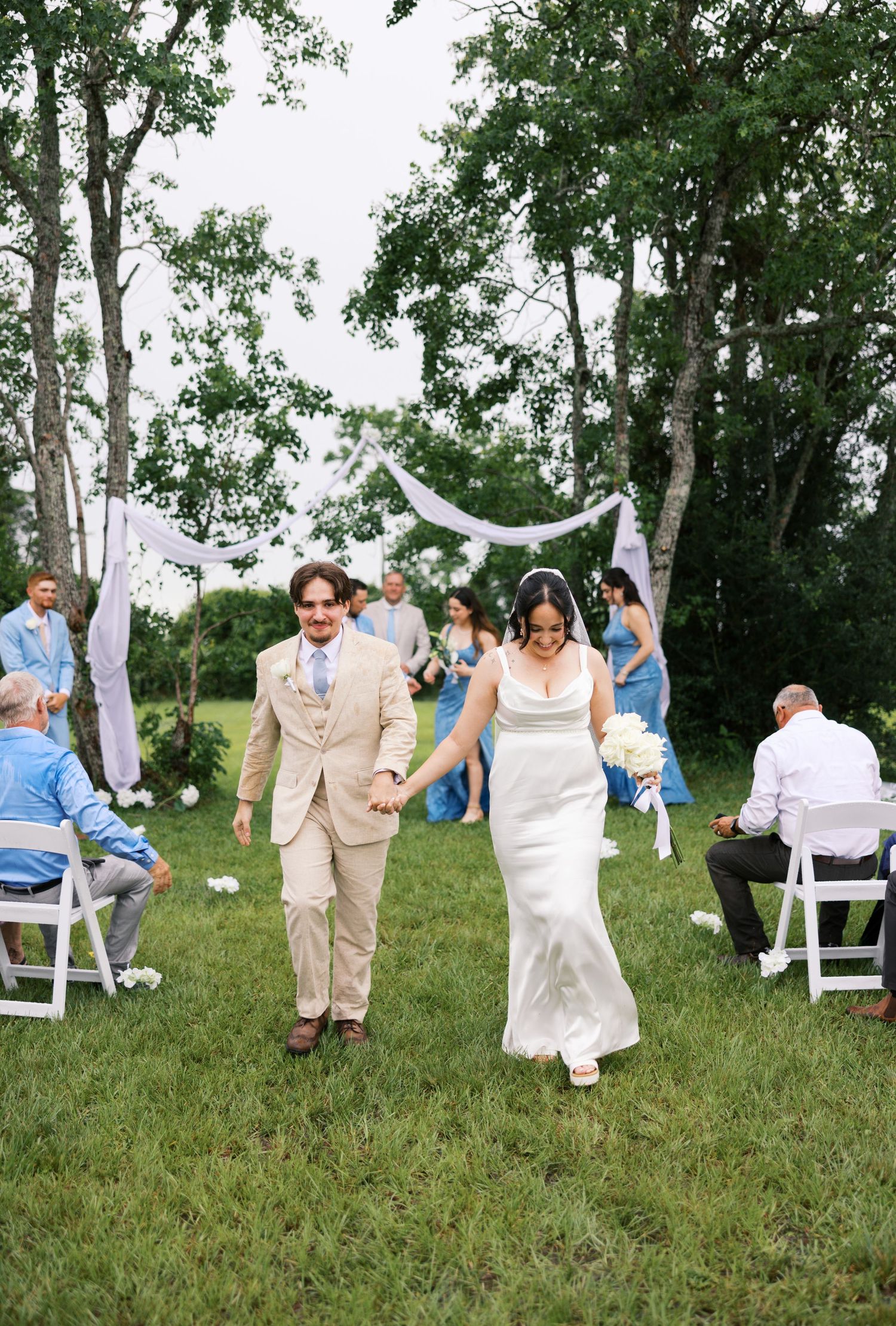 Bride and groom walk hand-in-hand down outdoor wedding aisle surrounded by guests in blue attire.