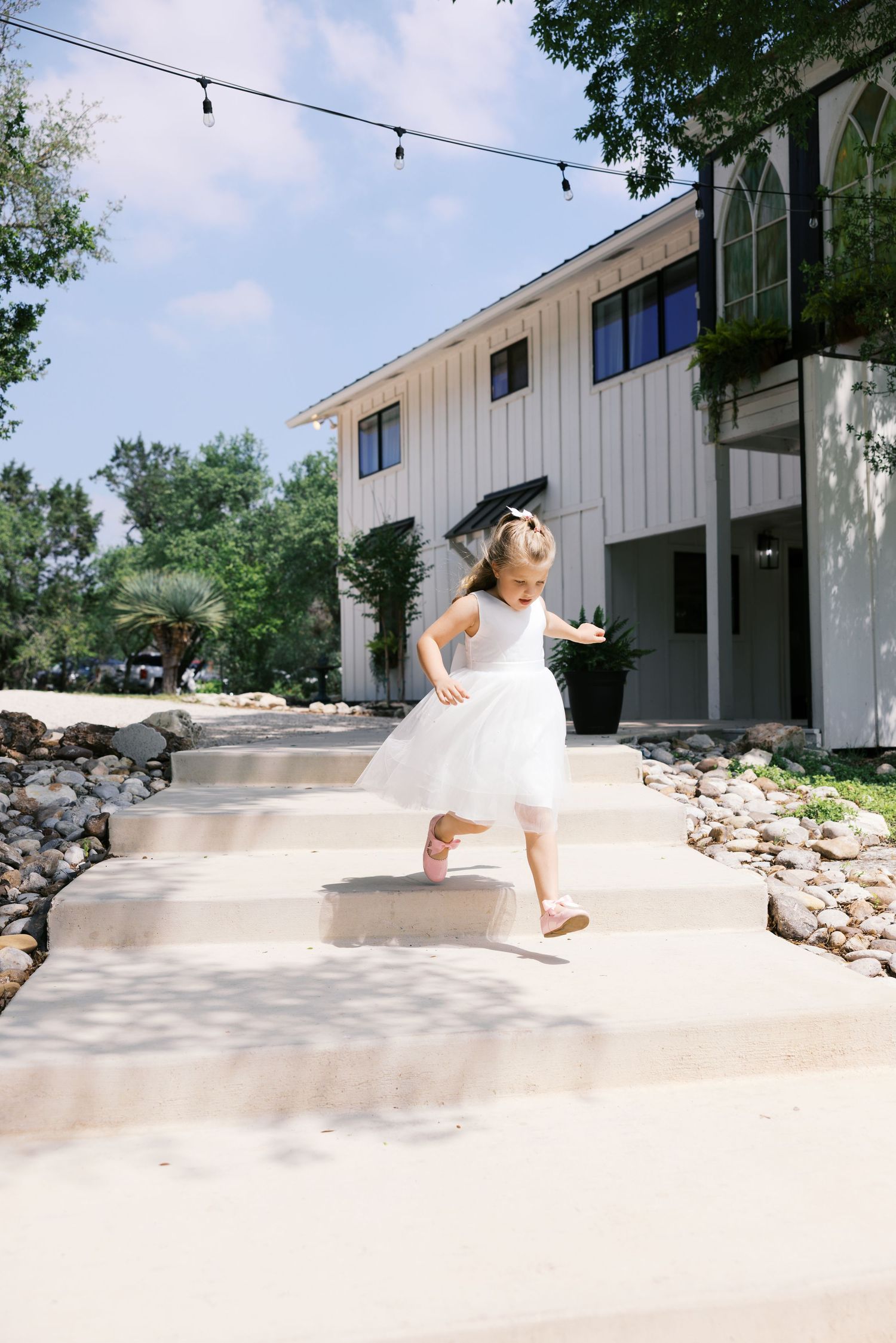 A little girl in a white tulle dress and graduation cap runs down concrete steps outside a white modern farmhouse.