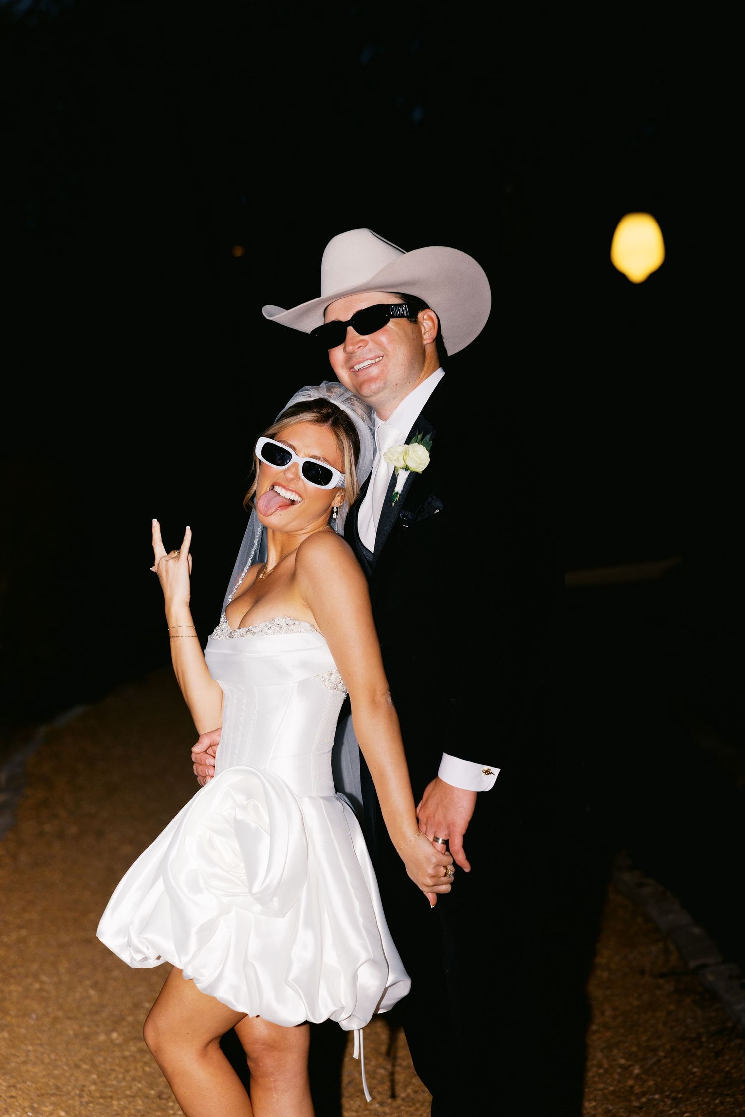 Bride and groom wearing sunglasses and cowboy hat at night, bride in short white dress making rock sign.