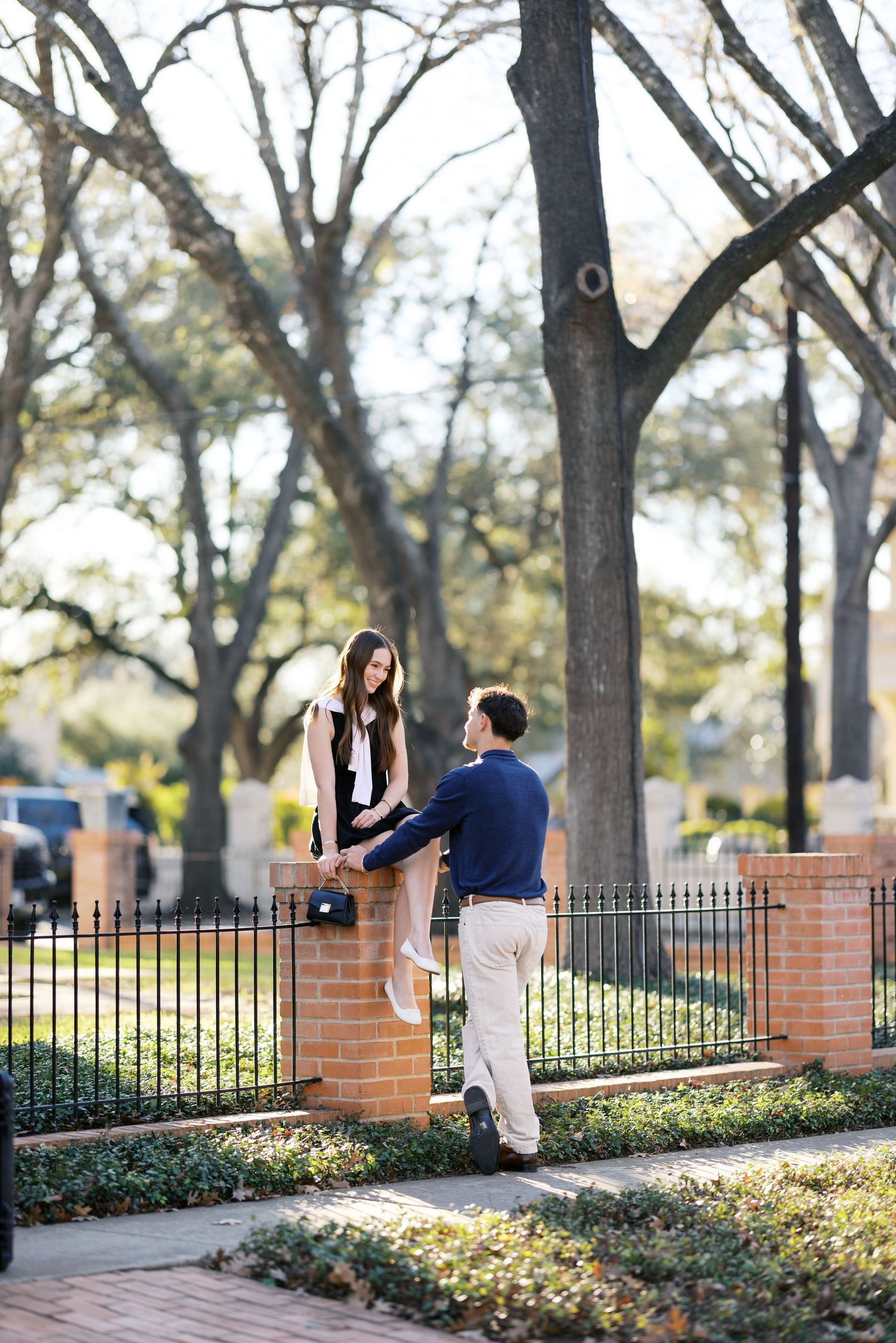 A young couple shares a romantic moment by a brick fence surrounded by trees.