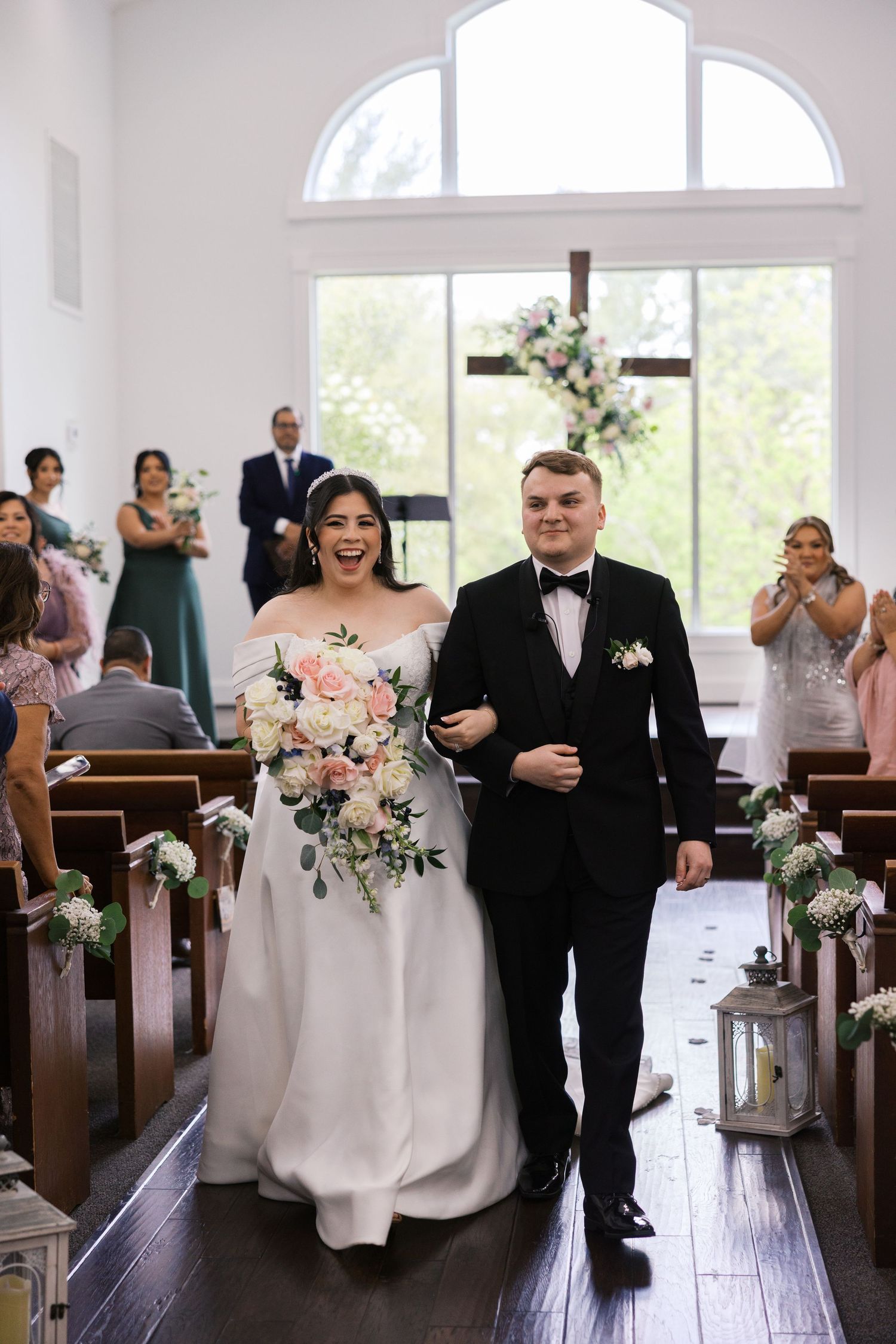 Bride and groom walk down the aisle of a white chapel, bride holding a blush bouquet, both smiling joyfully.