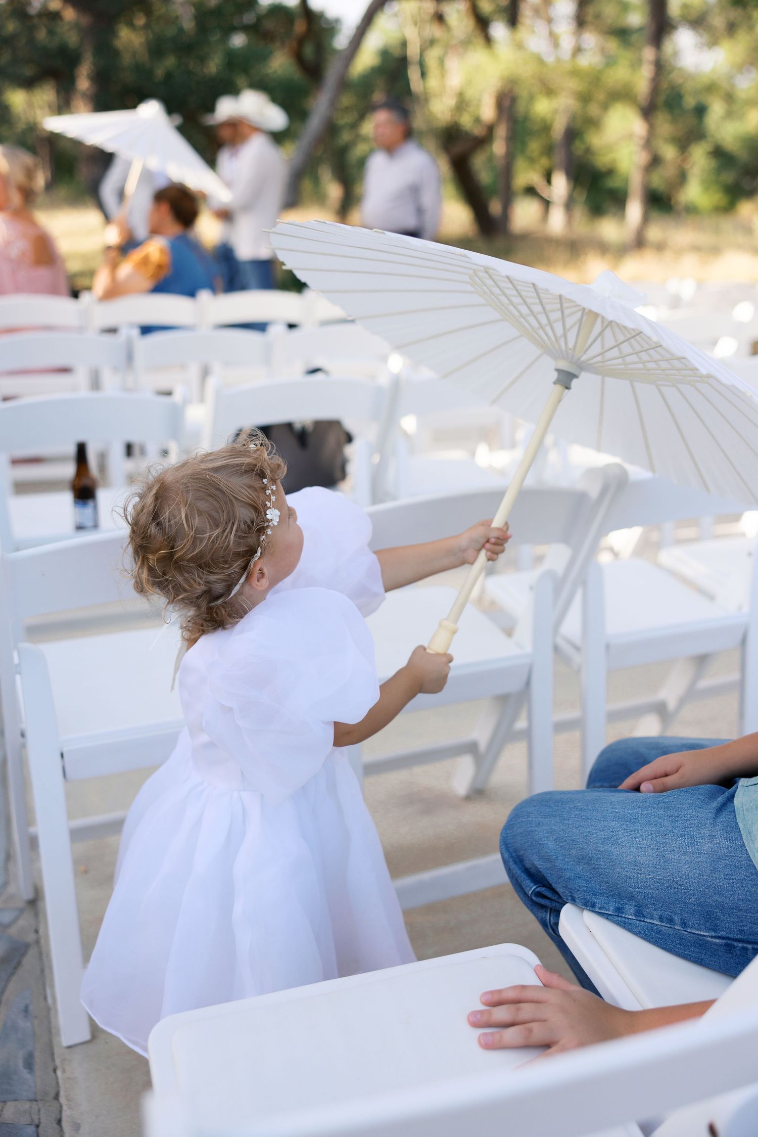 Small child in white dress holds decorative parasol among outdoor wedding chairs.