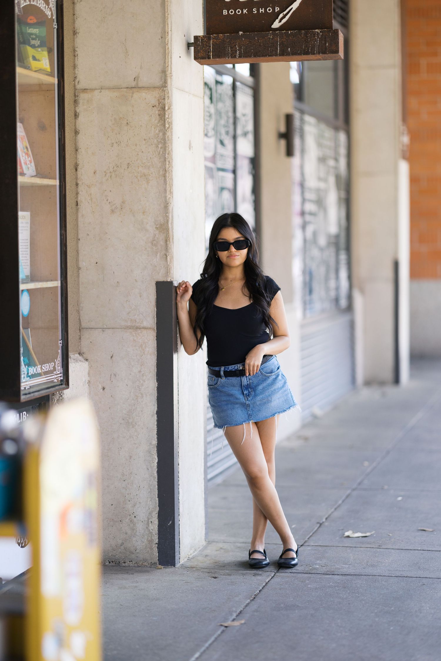 Woman in black top and denim shorts poses casually in front of a covered walkway with brick columns.