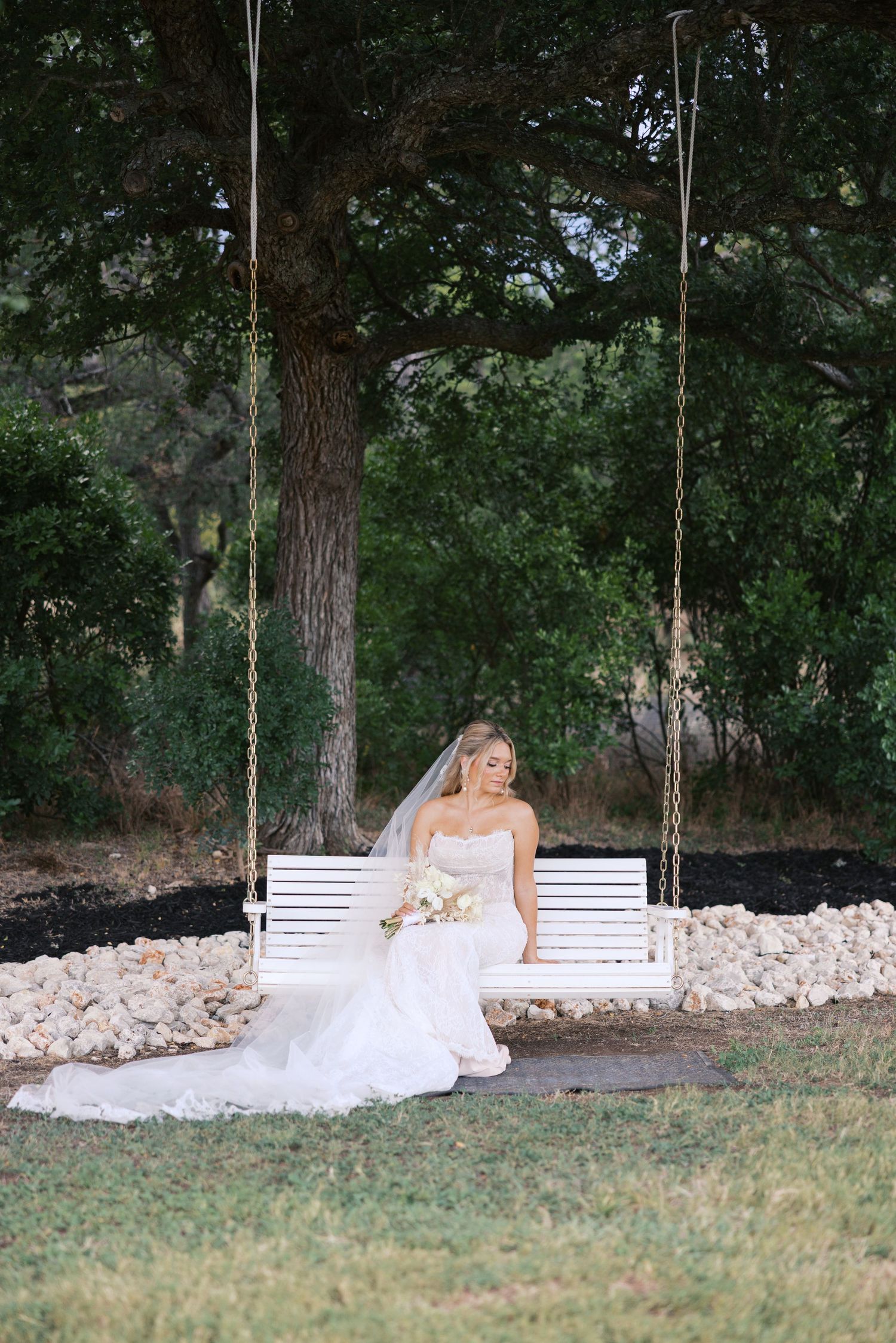 Elegant bridal portraits on white swing surrounded by trees and natural landscaping.