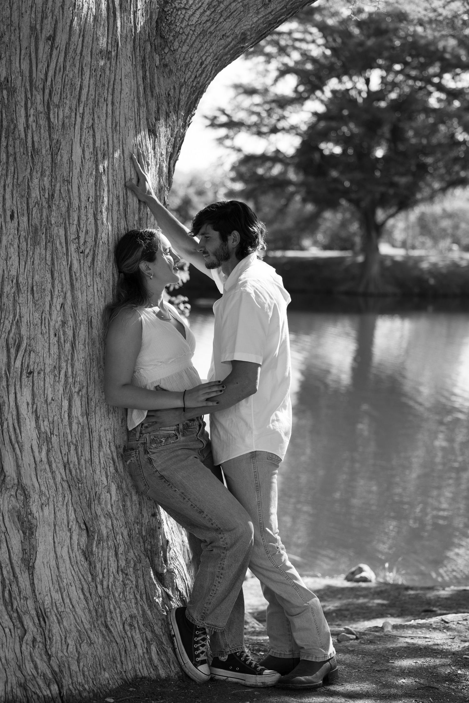 A black and white photo of a couple sharing an intimate moment against a large tree trunk by the water.